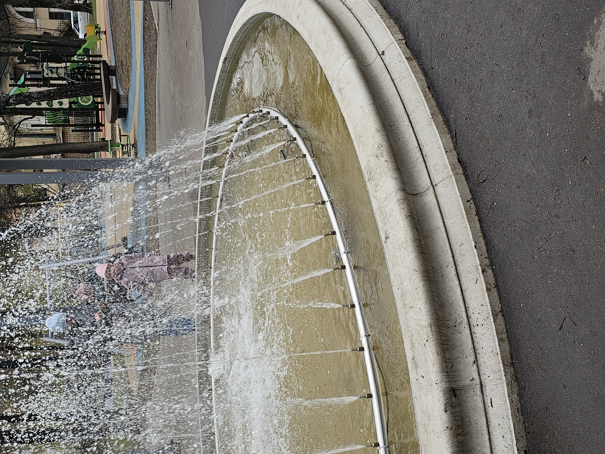 This image captures an outdoor scene featuring a curved fountain, a playground, and paved areas, set in what appears to be a park or public space in Khimki, Russia.
In the foreground, a large, semicircular fountain basin made of light-colored stone or concrete dominates the lower right portion of the frame. Water jets spray upwards and outwards from a metal pipe lining the inner edge of the basin, creating a significant amount of white spray. The water collected in the fountain's pool appears somewhat murky and yellowish-brown.
To the right of the fountain, a dark gray asphalt path or ground surface is visible, scattered with small bits of debris like leaves and twigs.
In the background, behind the fountain's spray, a vibrant playground area is visible. It features various pieces of green and brown equipment, including what looks like a spring rider shaped like a yellow creature, a slide, and possibly swings or other climbing structures. Several bare-branched trees are scattered throughout the background, suggesting either late autumn, winter, or early spring. Distant buildings are partially visible through the trees.
Several blurry figures, likely people, are positioned behind the fountain's water spray. One figure, possibly a child, wears a pink hat and a light-colored jacket, looking towards the fountain. Another figure is partially obscured nearby.
The scene depicts a daytime setting with soft lighting, possibly indicating an overcast day. The fountain is actively running, contributing to a lively atmosphere, while the playground suggests a recreational area. No visible text is present in the image.