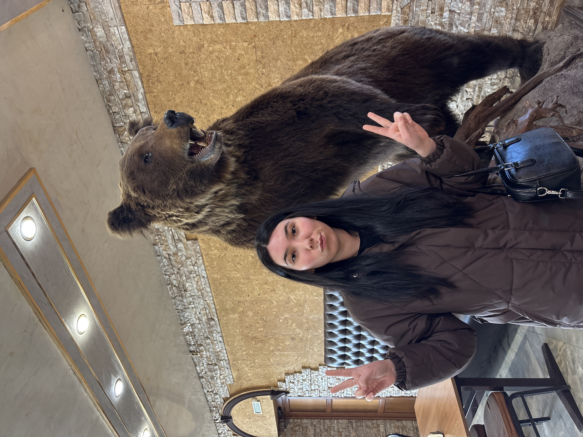A young woman poses next to a taxidermied brown bear in a restaurant in Mozhga, Russia. The woman has long dark hair, is wearing a brown puffer jacket, and is making a peace sign with both hands. The stuffed bear is mounted on a faux rock and tree branch display. The restaurant has a rustic aesthetic, with stone and faux wood paneling on the walls. Tables and chairs are visible in the foreground.