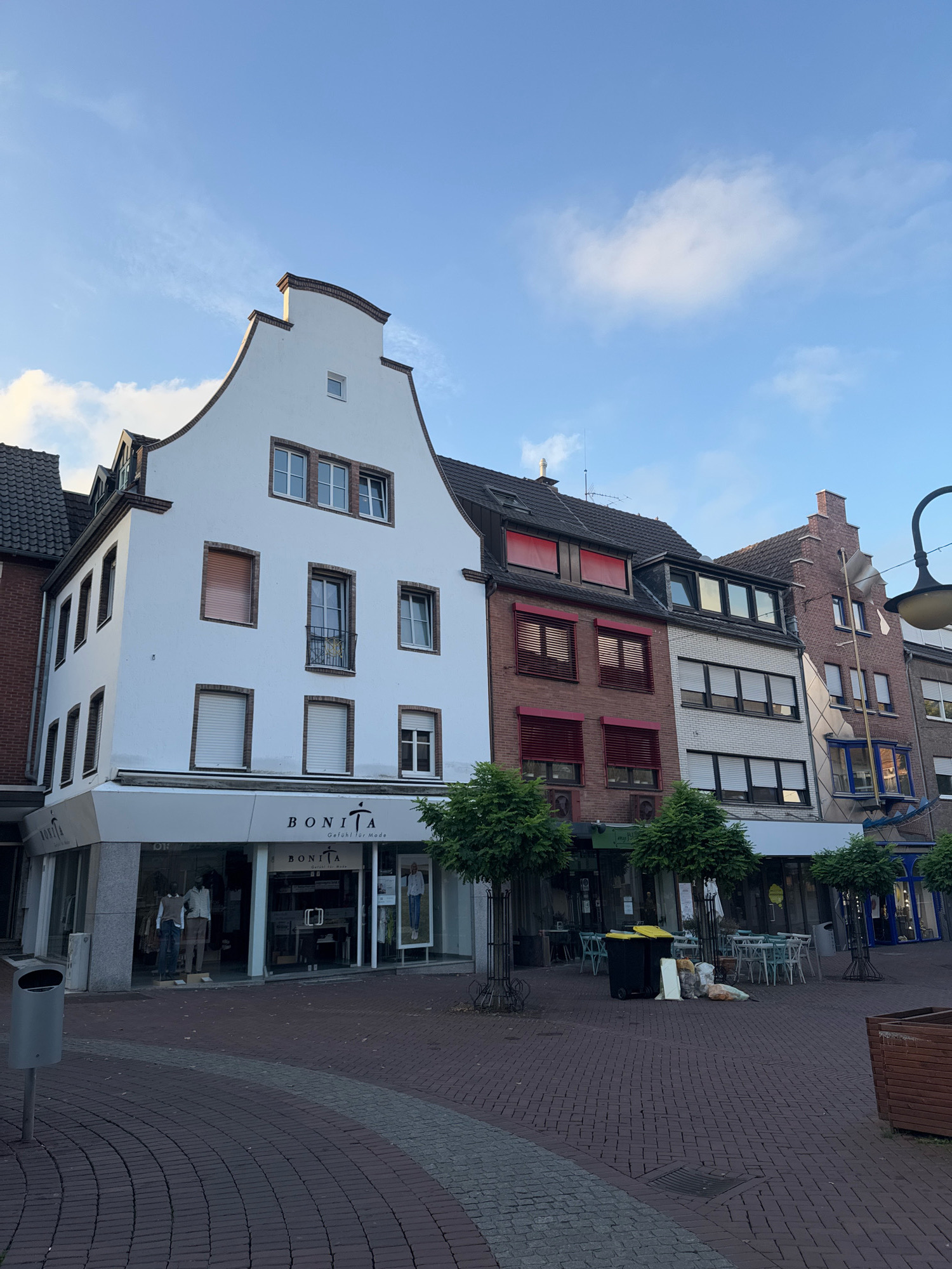 The image captures a street scene in Cleves, Germany, featuring a row of commercial buildings under a pale blue sky with scattered clouds. The architecture displays a mix of styles, with a prominent white building on the left featuring a stepped gable roof and brick accents around its windows. Below, the ground floor houses a "BONITA" clothing store with a large glass storefront displaying mannequins.
Adjacent to the white building is a red brick structure with multiple windows covered by red roller shutters. Further to the right, another building presents a white brick facade with dark-framed windows, also equipped with shutters. The southernmost building in the visible row is made of red brick and has a distinctive projecting bay window.
The street in front of the buildings is paved with reddish-brown brick, forming a pedestrian area. Several trees with rounded canopies are planted along the sidewalk, and outdoor seating with tables and chairs is arranged in front of some establishments. Trash receptacles, including a large yellow and black bin and a smaller grey bin, are visible, along with some scattered debris. A street lamp with a curved arm extends from the right side of the frame, partially obscuring the view of the buildings behind it. The overall impression is of a quiet, possibly early morning or late afternoon street in a European town.