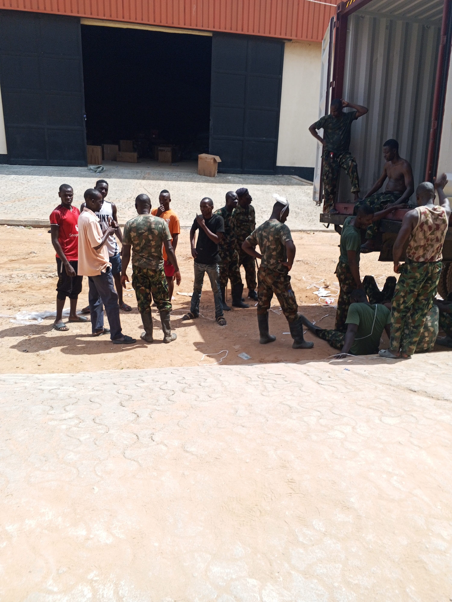 In Pasere, Nigeria, a group of men stand near a large building with an open loading bay, where several cardboard boxes are visible inside. Adjacent to the building is a freight container, where some men, clad in military camouflage, are seated and standing on the edge of the container. On the ground nearby, other men, some in military camouflage and some in civilian clothes, are gathered in a loose group. The ground is sandy and bears visible tire tracks. The sky is bright, casting harsh shadows.