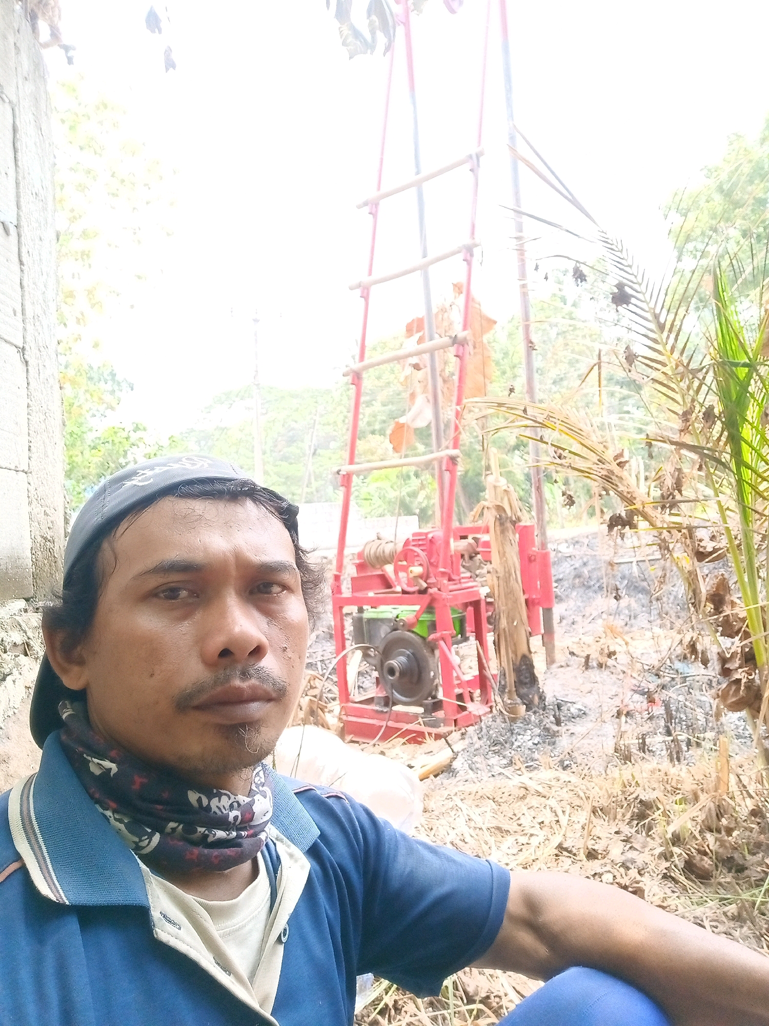 The image shows a selfie taken by a man in Banyuasih, Indonesia. The man is wearing a blue collared shirt over a white t-shirt, a dark baseball cap, and a patterned neck gaiter. He has a mustache and a goatee. In the background, a red drilling rig is set up, with bamboo poles extending upwards. The ground is covered in dry vegetation and what appears to be charred debris. Palm fronds are visible to the right of the frame. The overall environment suggests an outdoor, potentially rural or construction setting.