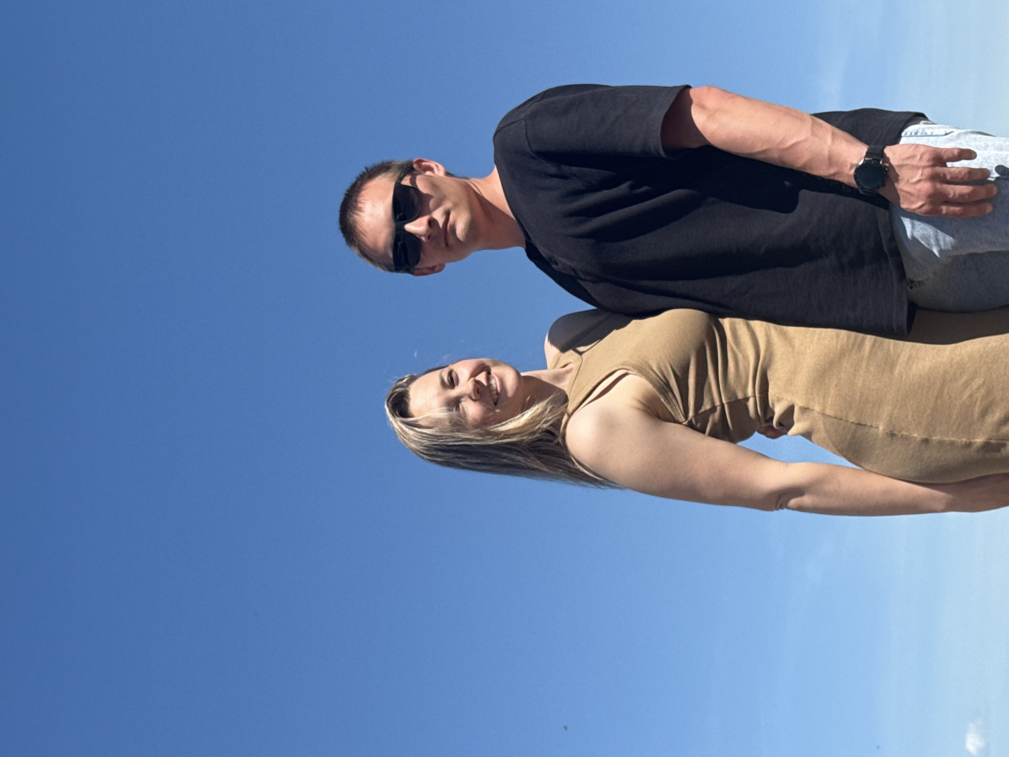 A low-angle shot features a fair-skinned man and woman against a clear blue sky. The man is wearing a black t-shirt, light-colored jeans, and dark sunglasses, and a watch on his left wrist. The woman is wearing a tan dress and has blonde hair. The exact location in the city of Chaikovsky, Russia is indiscernible.