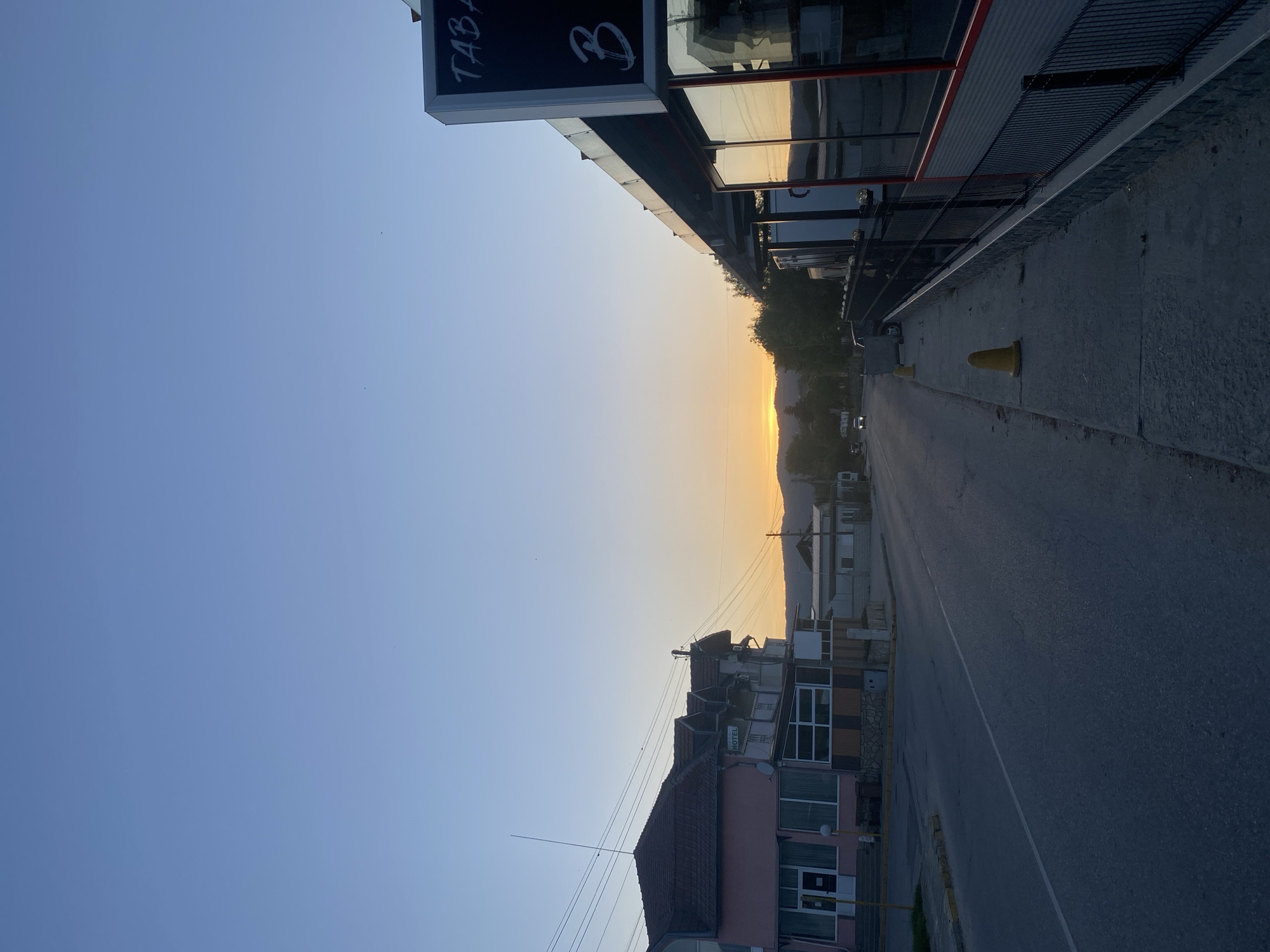 The image depicts a street scene in Satovcha, Bulgaria at sunset. The street runs between buildings, with the sky visible in the distance, exhibiting a gradient from blue to orange. On the left is a building with a sign that reads "TABA" followed by a Cyrillic letter. On the right is a building with reflective windows, a metal railing, and a concrete sidewalk. Traffic cones are positioned along the sidewalk. A street runs between these buildings. Buildings can be seen in the distance. Overhead power lines are visible.