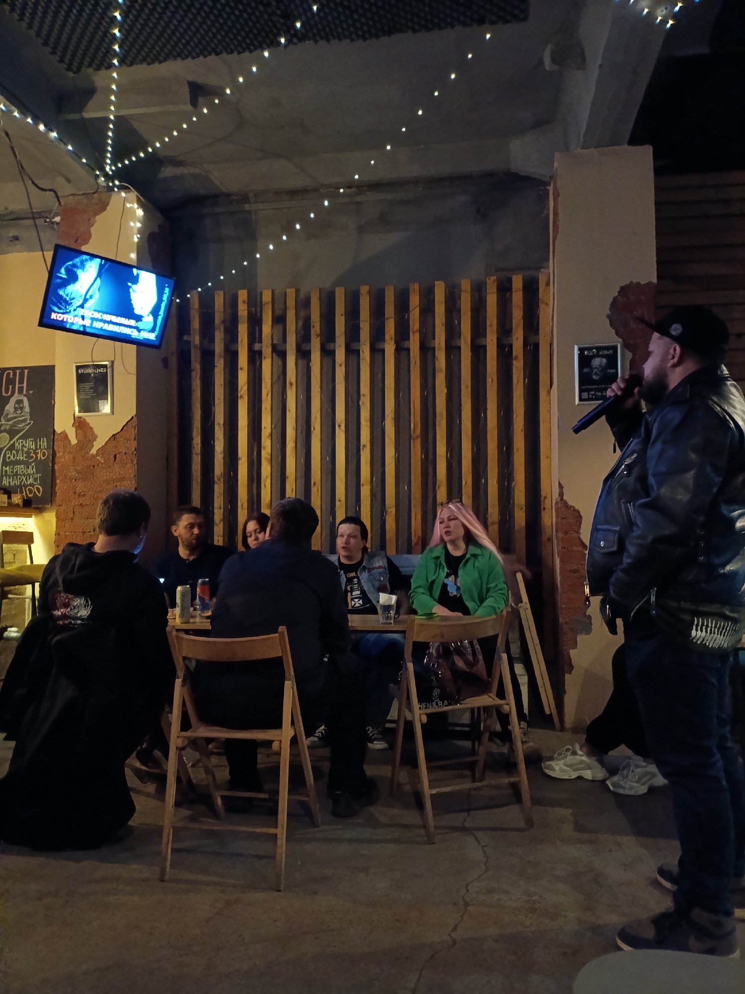 The image shows a group of people gathered at a table in what appears to be a bar or club in Chaikovsky, Russia. A person wearing a black leather jacket and a cap is standing and holding a microphone, seemingly giving a performance or speech. The other people are seated around the table, seemingly listening. There is a TV screen on the wall displaying some content, and some text and artwork on the wall as well. The environment appears to be dimly lit, with string lights hung across the ceiling. The atmosphere seems casual and social.