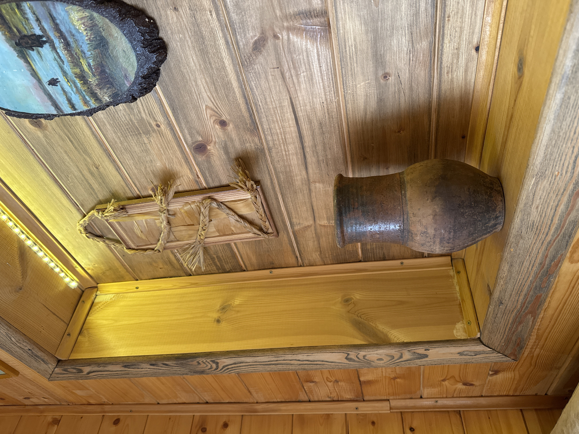 The image displays an upward view within a rustic interior, featuring extensive wooden paneling on the ceiling and walls. The wood exhibits varying light to medium brown tones with distinct grain patterns and visible knots, contributing to a natural, textured aesthetic.
Several decorative objects are integrated into this wooden setting. In the upper left quadrant, a round painting with a rough, bark-edged frame is mounted. It depicts a tranquil scene with light blue water and dark shapes, possibly animals such as bears, against a backdrop of trees and reflections. Below and to the right of the painting, a small, rectangular decorative piece, seemingly crafted from woven straw or raffia and tied with string, is attached to the wooden surface. Further to the right, a dark, aged earthenware jug or pot is placed on its side.
A recessed wooden shelf spans horizontally across the lower part of the image. Its interior is illuminated by a strip of LED lights visible on the left side, casting a warm, yellowish glow on the lighter wooden surfaces within the alcove. The front edge of this shelf features a darker wooden trim with pronounced grain patterns.
No people are present, and no particular activity is taking place; the scene is static, showcasing the decor of the space. The overall ambiance suggests a cozy, traditional, or cabin-like environment, consistent with a setting in Ново-Никольское, Russia. No visible text is present in the image.
