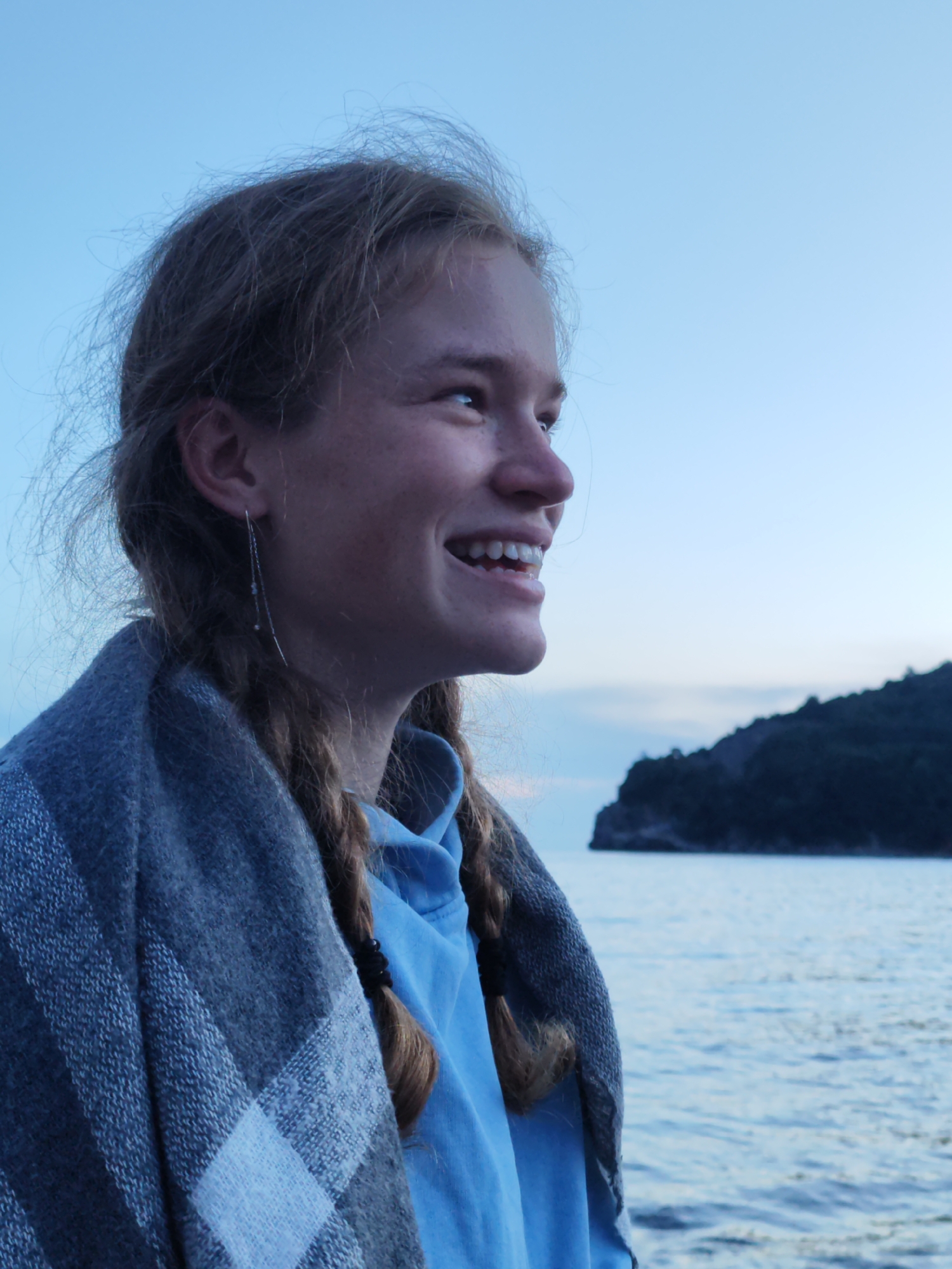 A portrait shot depicts a young woman with fair skin and hair, styled in braids, gazing to the right with a wide smile revealing her teeth. She is wrapped in a gray and blue plaid blanket and wears a blue sweatshirt. The background includes a body of water, and in the distance, a dark, hilly island or landmass is visible. The sky is a pale blue, suggesting either early morning or dusk. While the exact location within Budva cannot be determined from the image, the presence of water and a coastal land formation indicates a location along the Budva Riviera.