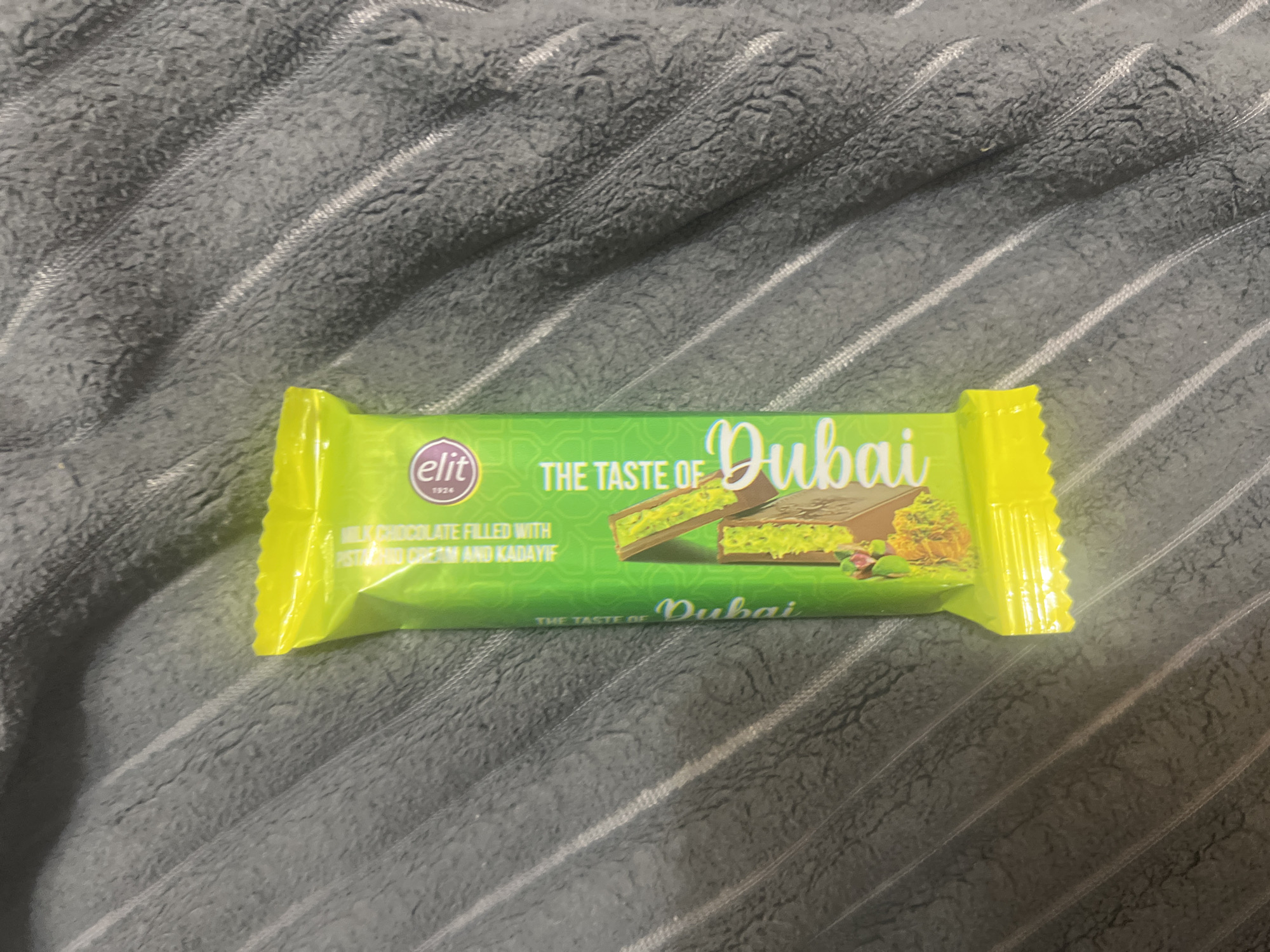 A bright yellow foil-wrapped chocolate bar with green lettering sits on a dark gray textured blanket. The chocolate bar has an image of a broken chocolate bar with green filling and some nuts. The lettering on the chocolate bar reads "ELIT" in a circle with "1926" below it. Below that it says "MILK CHOCOLATE FILLED WITH PISTACHIO AND KADAİF". Further down the bar, the text "THE TASTE OF Dubai" is visible in white cursive lettering. The chocolate bar is positioned diagonally on the blanket. The blanket appears to be a soft, fuzzy material with raised ridges running diagonally across it. The scene is indoors, with no discernible background or context of its location in Zheleznogorsk, Russia.