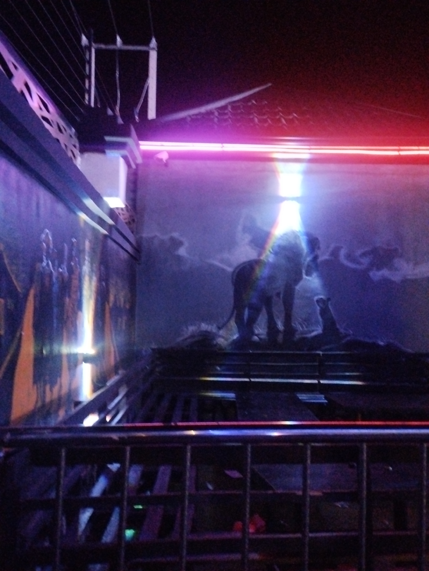 The image shows a dark, illuminated outdoor scene at night. A mural depicting an elephant standing on what appears to be a raised platform dominates the background. The mural is lit with bright, slightly diffused light. The mural's artwork has clouds in the background. A wall to the left is also decorated with artwork, including what appears to be human figures.
A metal railing is visible in the foreground, obscuring the view of the space directly in front of the mural. Above, there are electrical wires. A red neon light is visible above the mural. The location is in Tumfure, Nigeria.