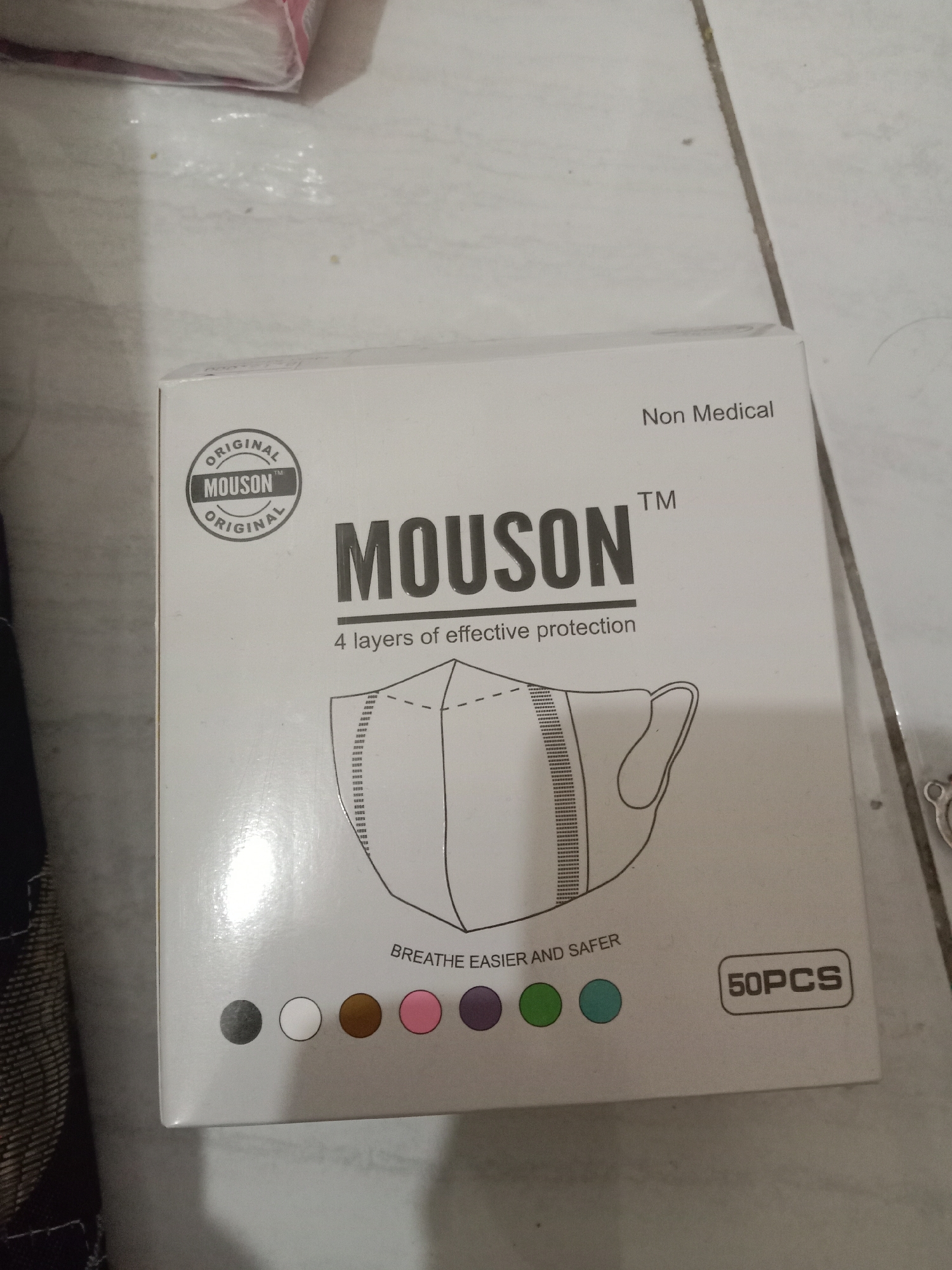 The image shows a retail box of Mouson brand masks, labeled "4 layers of effective protection." The box also states "Non Medical" and "50PCS". Below the mask illustration are circles representing different color options for the masks. The box is placed on a floor with a tiled or patterned surface. A portion of what appears to be a package of tissues is visible in the top left corner. The lighting suggests an indoor setting. There is no indication of the city of Brangsong, Indonesia within the image itself.