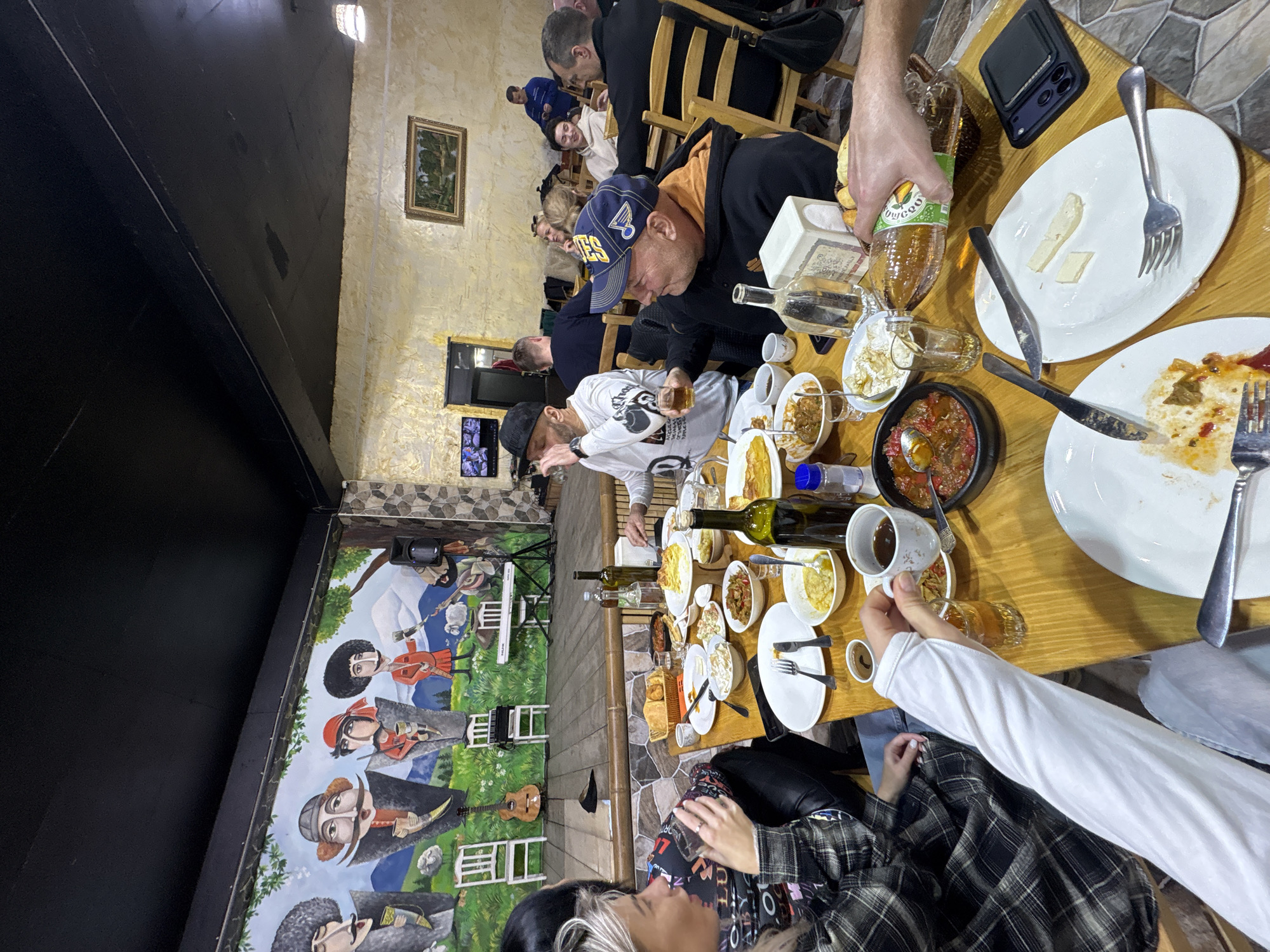 A group of people are gathered around a wooden table laden with food and beverages in what appears to be a restaurant. The table holds plates, bowls, glasses, silverware, and bottles, suggesting a meal is in progress. Several individuals are visible around the table, some seated on wooden chairs. One person is wearing a cap with "Blues" visible on it. Another person wears a black cap and a white t-shirt.
In the background, a wall mural depicts figures in historical or traditional attire. The restaurant's interior features stone-like wall patterns and dim lighting. A painting hangs on the wall above the people in the background. Based on the prompt, this scene could be taking place in Kveda Chkhutuneti, Georgia.