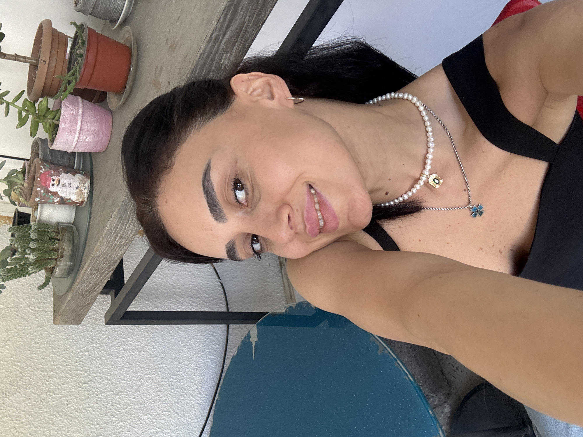 A woman is taking a selfie outdoors, with a shelf of potted plants in the background. She has dark hair, brown eyes, and is wearing a black top and a pearl necklace with two pendants. The plants are on a wooden shelf supported by a black metal frame. The scene appears to be in a bright, natural light, suggesting it is daytime. The woman's expression is relaxed and she is smiling slightly. The backdrop includes a textured white wall and a blue surface, possibly a table or a floor. The location is specified as Tbilisi, Georgia.