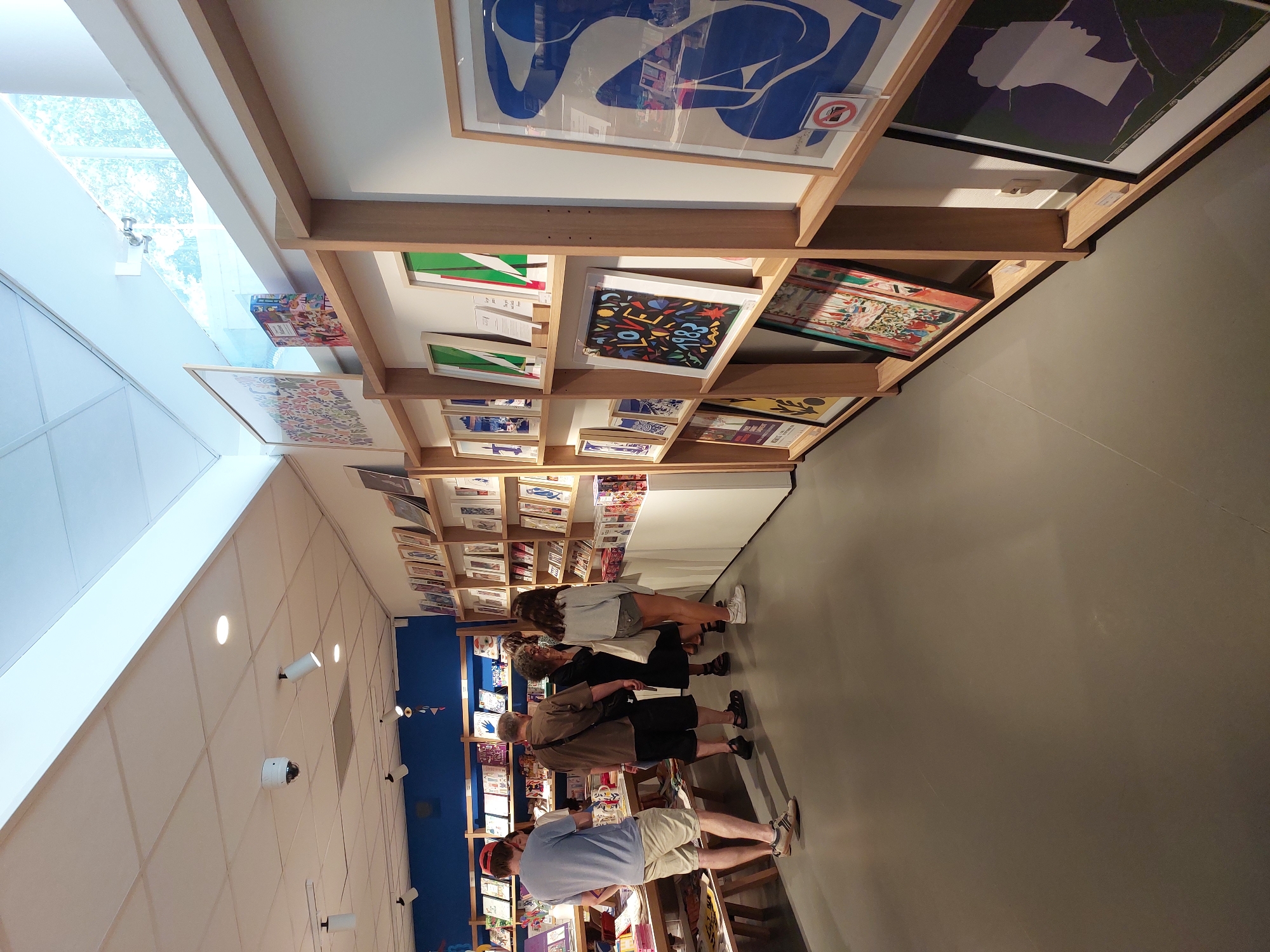 The image shows the interior of a shop with a blue wall in the background and shelves displaying artwork. Several people are browsing the merchandise. The shop appears to be a bookstore or gallery, with books and framed prints on display. The flooring is a smooth, grey material. The lighting is bright, with ceiling lights and natural light coming from a window at the top of the image. One person is wearing a red cap and a blue t-shirt, another is wearing a brown shirt and black shorts, and a third is wearing a white t-shirt and denim shorts. There is a large framed blue artwork on the right side of the image. The environment suggests a retail space designed for exhibiting and selling art.
