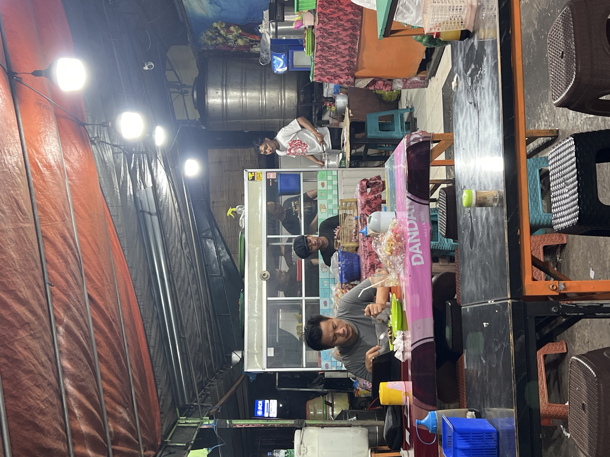 This image captures a nighttime scene in Denpasar, Indonesia. An outdoor food stall is active with several people present.
Three men are visible behind the stall's counter, likely preparing or serving food. One man wears a gray shirt and appears to be interacting with something on the counter. Another man, wearing a black shirt and hat, stands behind him. A third man in a white shirt is further back, seemingly engaged in food preparation.
The stall is constructed with metal framing and has some see-through sections with window. The surrounding environment includes a tarpaulin-covered structure on the left, illuminated by bright light bulbs. To the right, tables and chairs are set up for customers. Various objects such as water bottles, containers, and cooking utensils are scattered on and around the stall's counter. A BCA bank logo is seen.