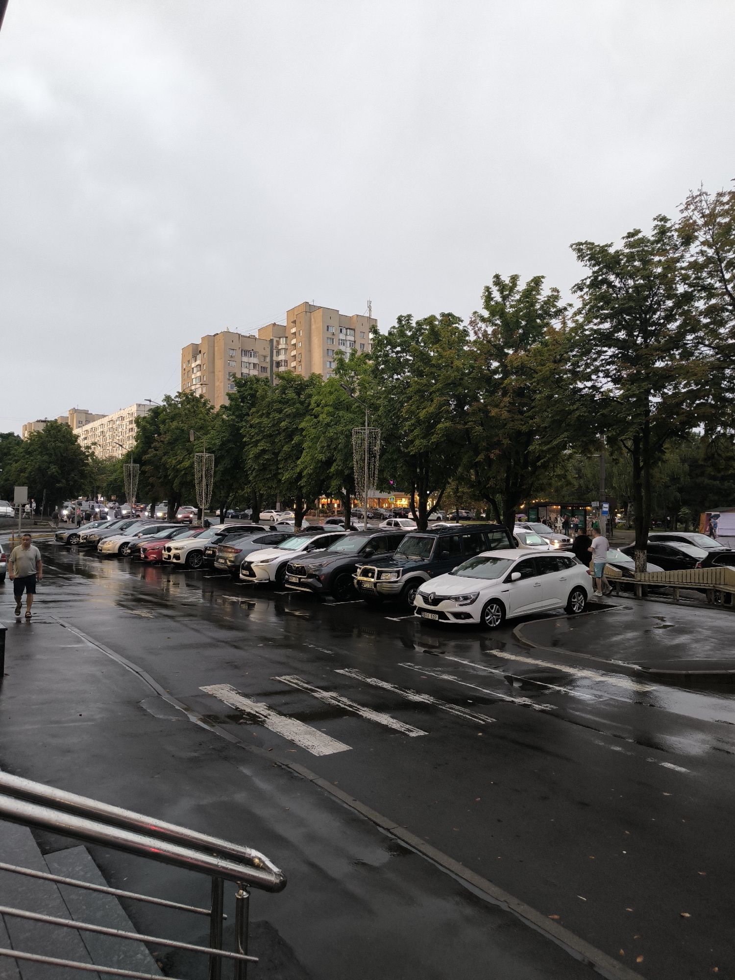 The image depicts a wet street scene in Chișinău, Moldova. The sky is overcast, suggesting a cloudy or rainy day. A row of cars is parked along the street. The wet asphalt reflects the lights of the vehicles and street lamps. In the foreground, a man wearing a light-colored t-shirt and dark shorts walks on the sidewalk. Further down the street, several buildings with multiple stories are visible. Trees with green foliage line the street between the buildings and the parking area. Decorative lights, possibly for an event, are strung between poles along the sidewalk. The overall scene conveys an urban environment with a focus on the parked vehicles and pedestrian activity.