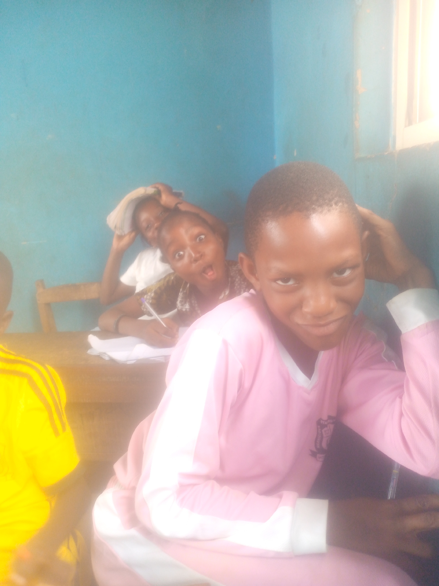The image shows a classroom scene in Unknown, Unknown. Three children are visible. One child is in the foreground, wearing a pink shirt with a white collar and sleeve trim. The child is leaning against the table and looking at the camera. Behind the child, two other children are sitting at a wooden desk and writing on paper. One is wearing a patterned brown shirt. The third child is holding papers over their head and is wearing a white shirt. In the foreground, the left side of a fourth child in a yellow shirt is barely visible. The walls of the classroom are blue. A window is on the right of the frame.