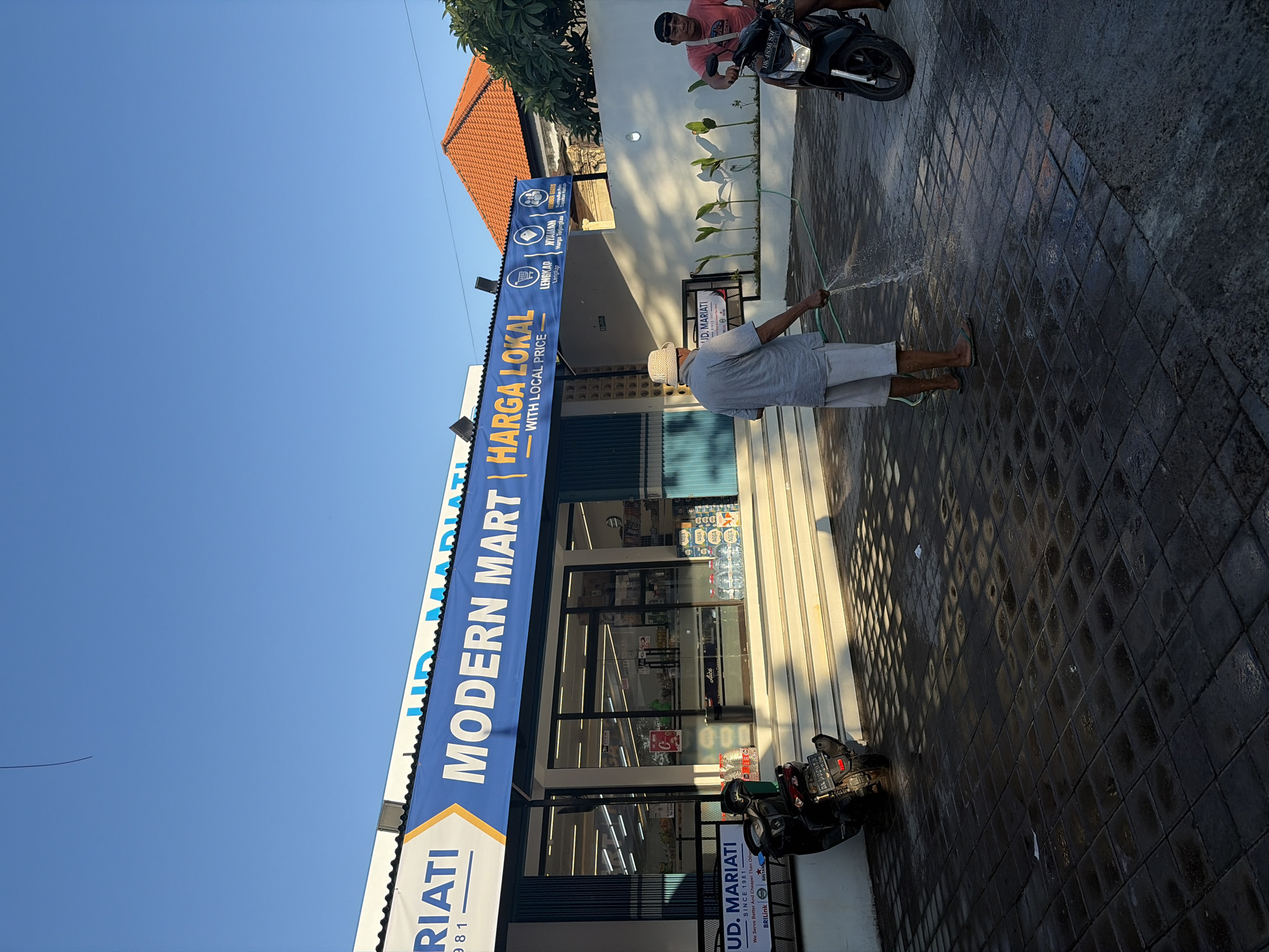 The image depicts the exterior of a "MODERN MART" store in Kubu, Indonesia. The store has a blue awning with white lettering and a yellow accent. In front of the store, two men are visible. One man, wearing a hat and light-colored shorts, is using a green hose to spray water on the paved ground. The other man, wearing a red t-shirt, is sitting on a motorcycle parked to the right. A black motorcycle is parked to the left of the store entrance. The ground is paved with dark square tiles, and there are shadows cast by the sunlight. The sky is clear and blue. The building has large glass windows revealing products inside. A sign on the left side of the entrance reads "UD. MARIATI SINCE 1991".