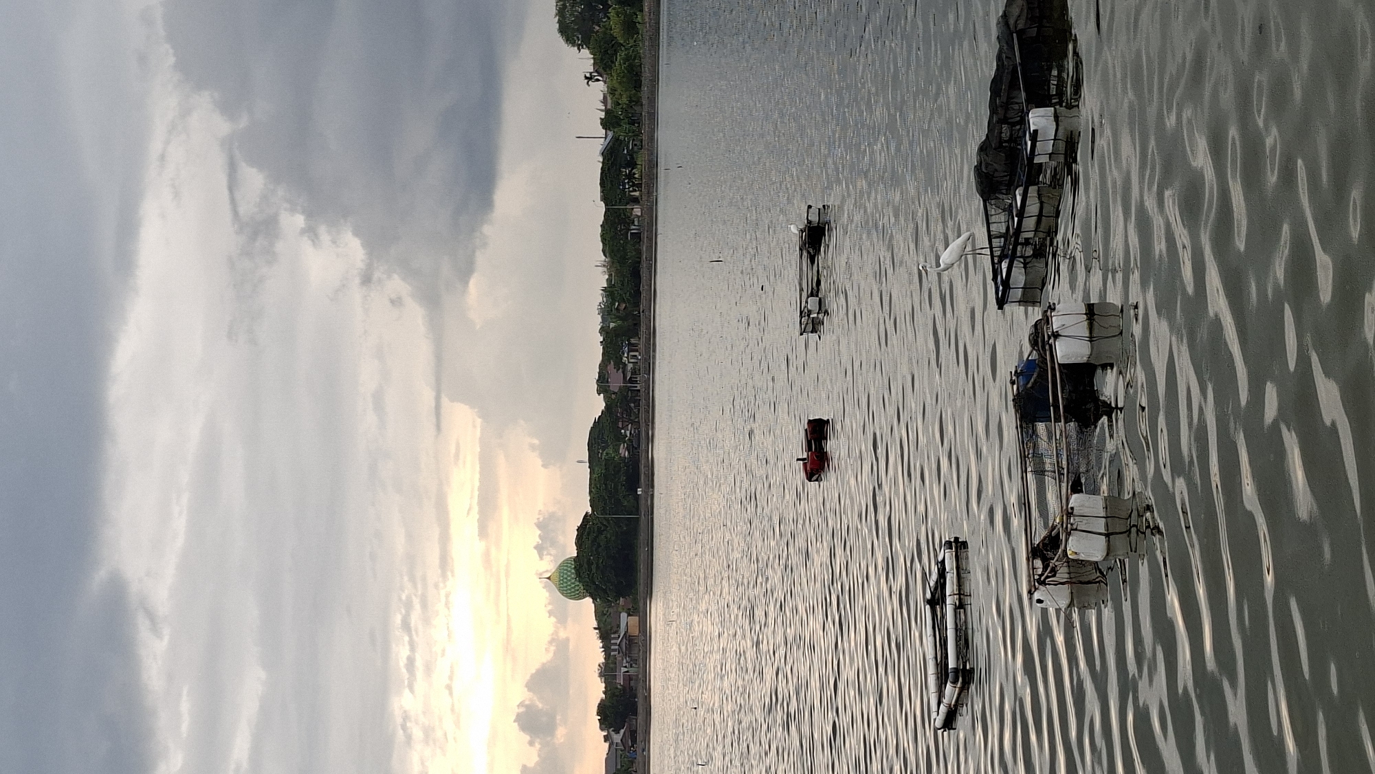 This is a photograph of a body of water, likely a lake or reservoir, in the city of Lhokseumawe, Indonesia. The sky is overcast with a soft light suggesting it is either sunrise or sunset. In the background, a green domed building, possibly a mosque, is visible, surrounded by trees and some low-rise buildings. The water's surface is rippled, reflecting the cloudy sky. Scattered across the water are several floating structures, likely fish cages or platforms, constructed with white buoys and dark netting. One white bird, possibly an egret or heron, stands on one of these platforms in the mid-ground, and another appears in the lower right of the frame. A small red object, possibly a buouy or small boat, is also visible floating on the water. The overall scene depicts a calm, natural environment within an urban setting.