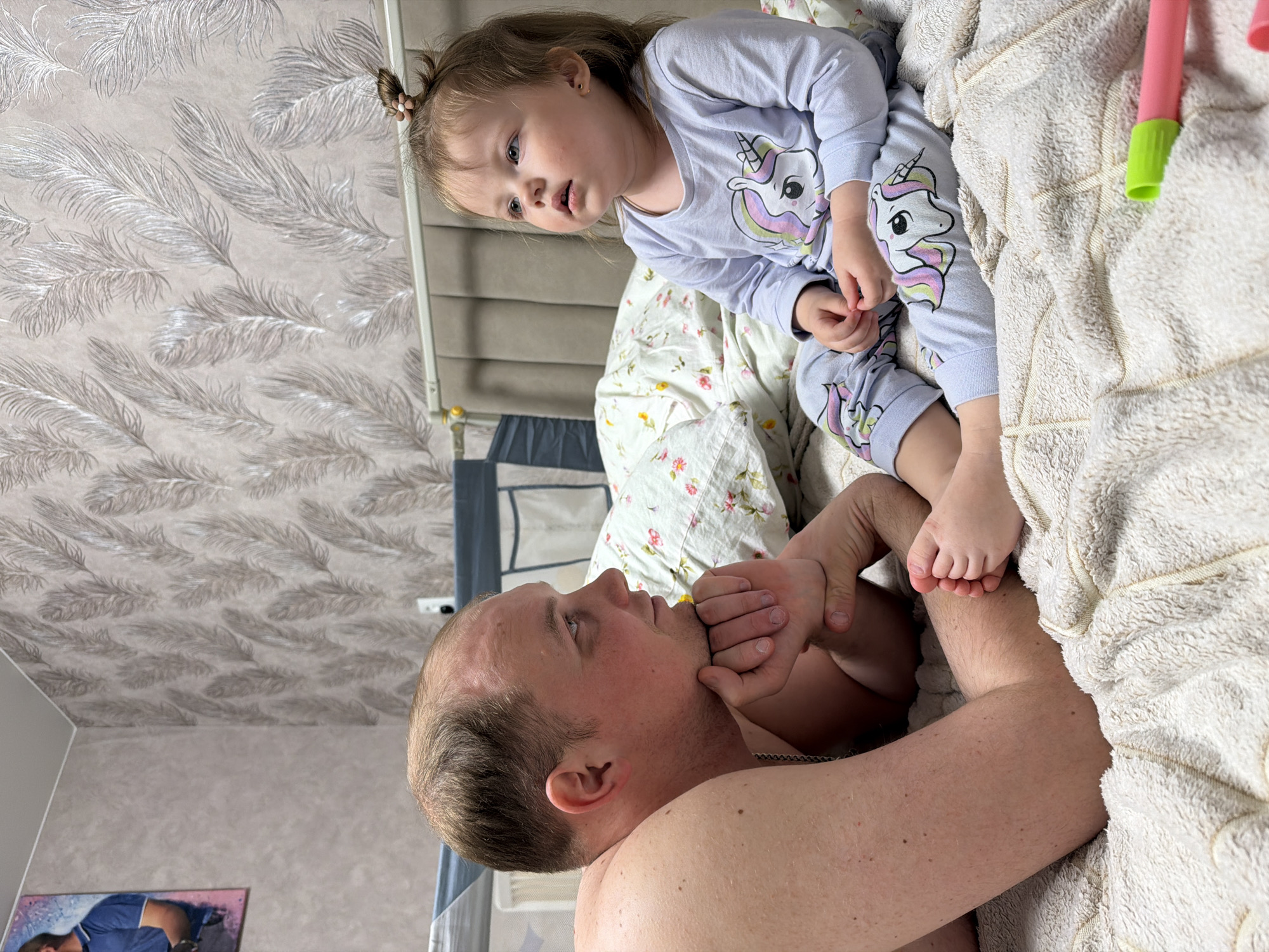The image captures an intimate moment between a man and a young child in a home setting in Reshetnikovo, Russia. The man, with short, light brown hair and a visible chain necklace, is shirtless and appears to be lying on his back, looking upwards. His hands are near his head. The child, likely a girl, has light brown hair styled in two small pigtails with flower-shaped hair ties. She is wearing light purple pajamas adorned with unicorn prints. She is positioned above the man, possibly sitting on the edge of a bed, looking down at him with a curious or focused expression. Her small feet are resting playfully on the man's chest or shoulder, and her hands are gently holding his face or chin.
The setting is a bedroom, indicated by the bed with white and floral patterned bedding, and a plush, cream-colored blanket draped over part of it. The wallpaper in the background features a distinctive silver feather pattern against a light grey or beige wall. A small pink and green toy or object is partially visible on the blanket near the child. A framed picture, too blurry to discern details, is on the wall in the lower left background, along with what appears to be a security camera or light fixture. The interaction suggests a loving, playful, and tender familial bond, possibly during morning or evening hours given the pajamas. There is no discernible text visible in the image.