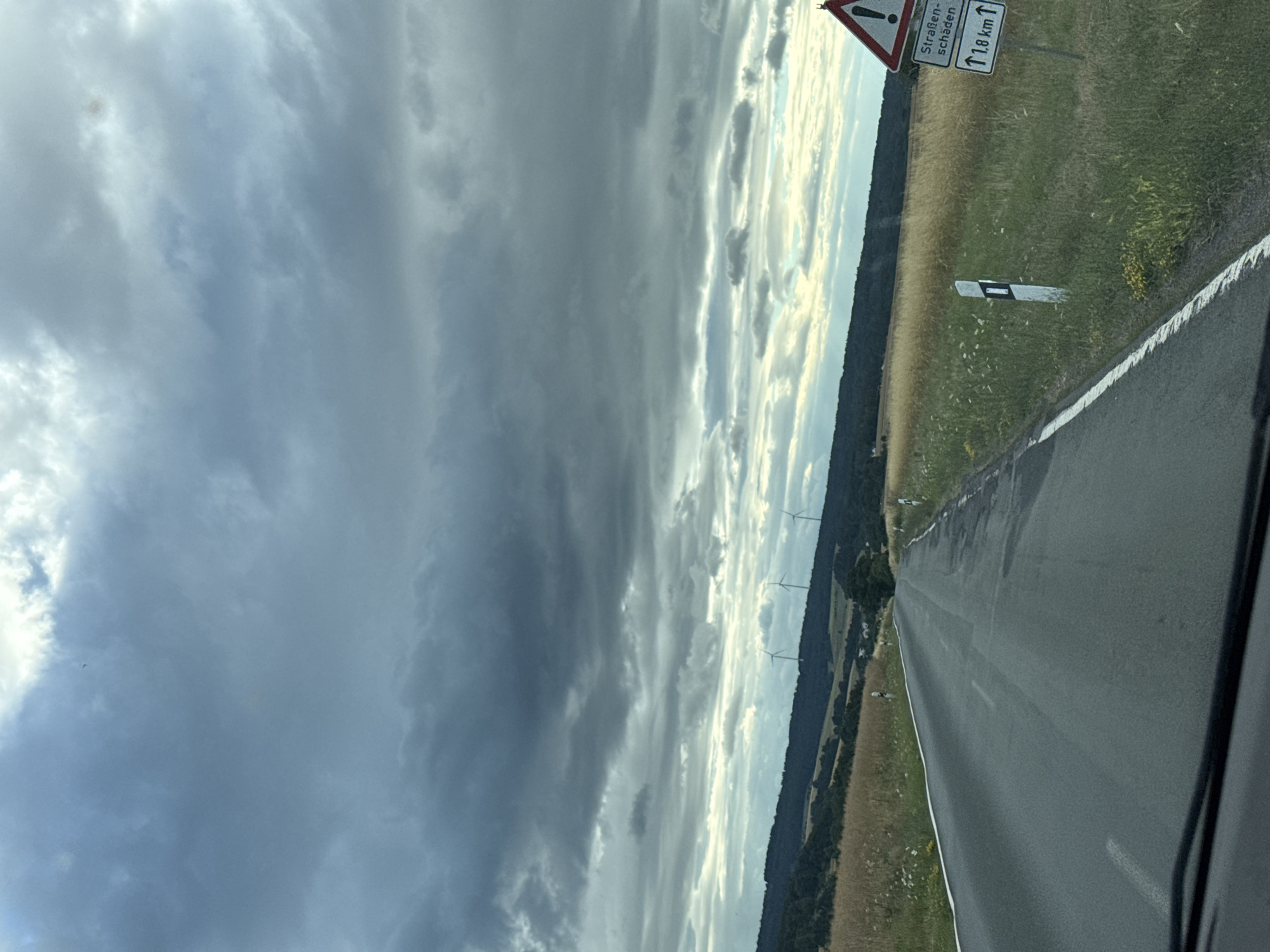 The image depicts a road in Oberwetz, Germany, viewed from a car. The sky is partly cloudy, with a mix of dark, dramatic clouds and brighter, sunlit patches, suggesting a dynamic weather pattern. The time of day appears to be late afternoon or early evening, given the low angle of the sun.
The scene shows a two-lane asphalt road stretching into the distance, bordered by grassy fields and a treeline on the right. On the left side of the road, there are several wind turbines visible in the middle distance, indicating a rural or semi-rural landscape. A white road marker with black markings is present in the field to the right of the road.
Attached to a post on the right side of the road are two German traffic signs. The top sign is a triangular warning sign with a red border and an exclamation mark, indicating a general hazard. Below it, a rectangular sign has text in German and an arrow pointing upwards. The text reads "Straßen- schaden" and "1,8 km". This translates to "Road damage" and indicates that the road damage is 1.8 kilometers ahead.
The overall mood is somewhat dramatic due to the contrast between the dark clouds and the sunlight. The image captures a moment of travel on a German road, with visual cues of both natural elements and man-made infrastructure.