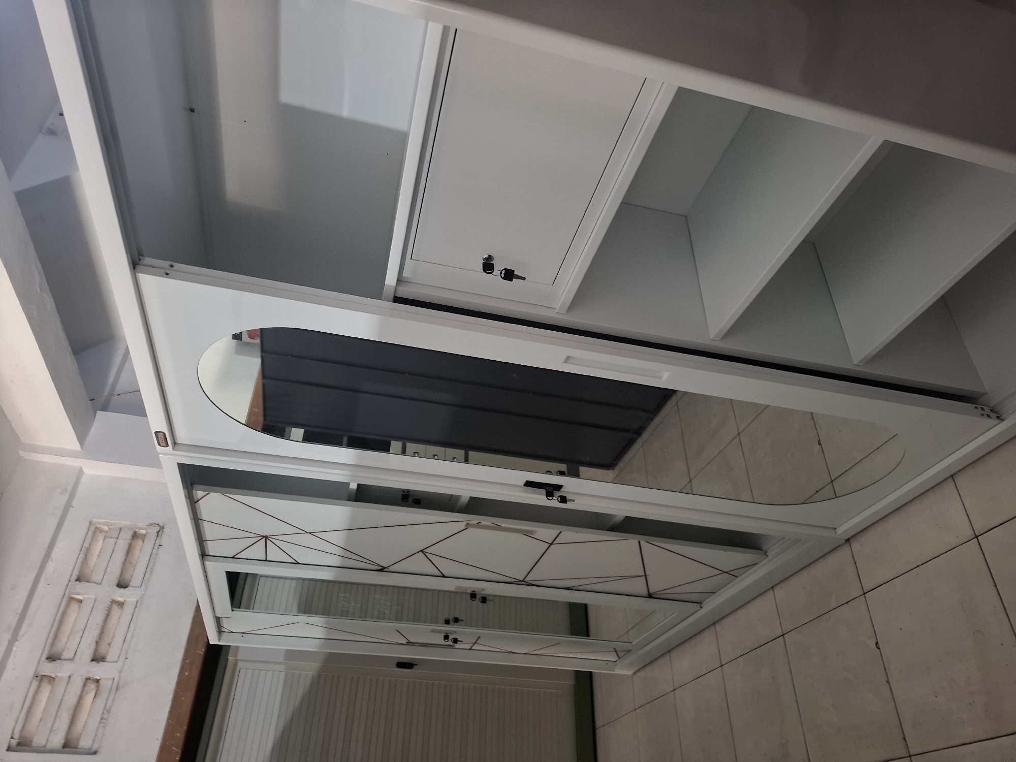The image shows a white, modern closet with mirrored doors that have a geometric pattern. The closet is in a room with tiled floors and a vent on the wall. The closet appears to be new and unused. There are no people in the image. The room is dimly lit, with the primary light source appearing to be coming from the left side of the frame, possibly from a window or door that is not fully visible. The overall impression is that this is a retail display or a newly furnished room.