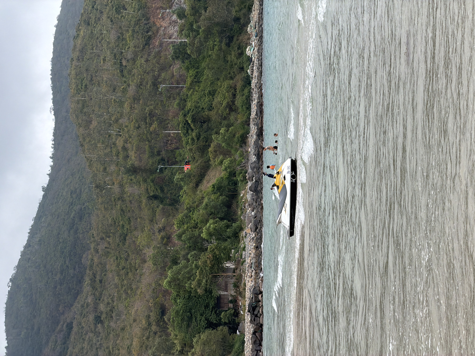 The image depicts a coastal scene in Nam Nha Trang Ward, Vietnam. A forested mountain slopes down to a rocky shoreline where a yellow and black personal watercraft (PWC) sits partially in the water, with several individuals standing nearby. Two other people are in the water, suggesting recreational activity. The water is grey and choppy. The sky is overcast, contributing to the overall muted color palette. The vegetation on the mountainside is dense and varied. In the background, there is a red flag on a utility pole.