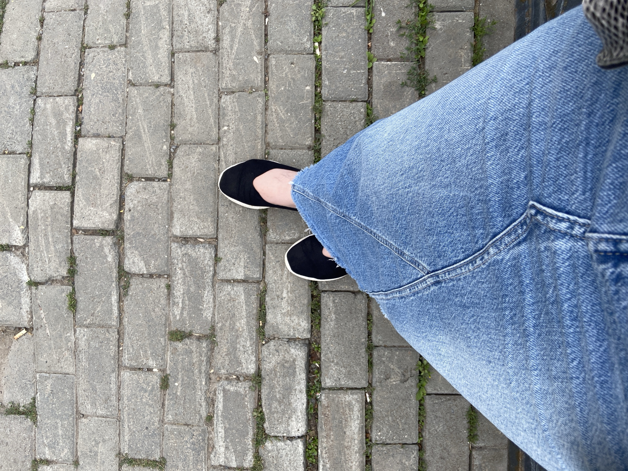 The image shows a person's legs and feet from a top-down perspective, standing on a paved walkway. The walkway is made of rectangular gray stone blocks with patches of green grass growing in the cracks. The person is wearing blue denim jeans that appear slightly faded. On their feet, they have black slip-on shoes with white soles. The lighting suggests it is daytime, and the scene appears to be outdoors in an urban or park setting. There are no visible text elements in the image. The overall impression is casual and everyday.