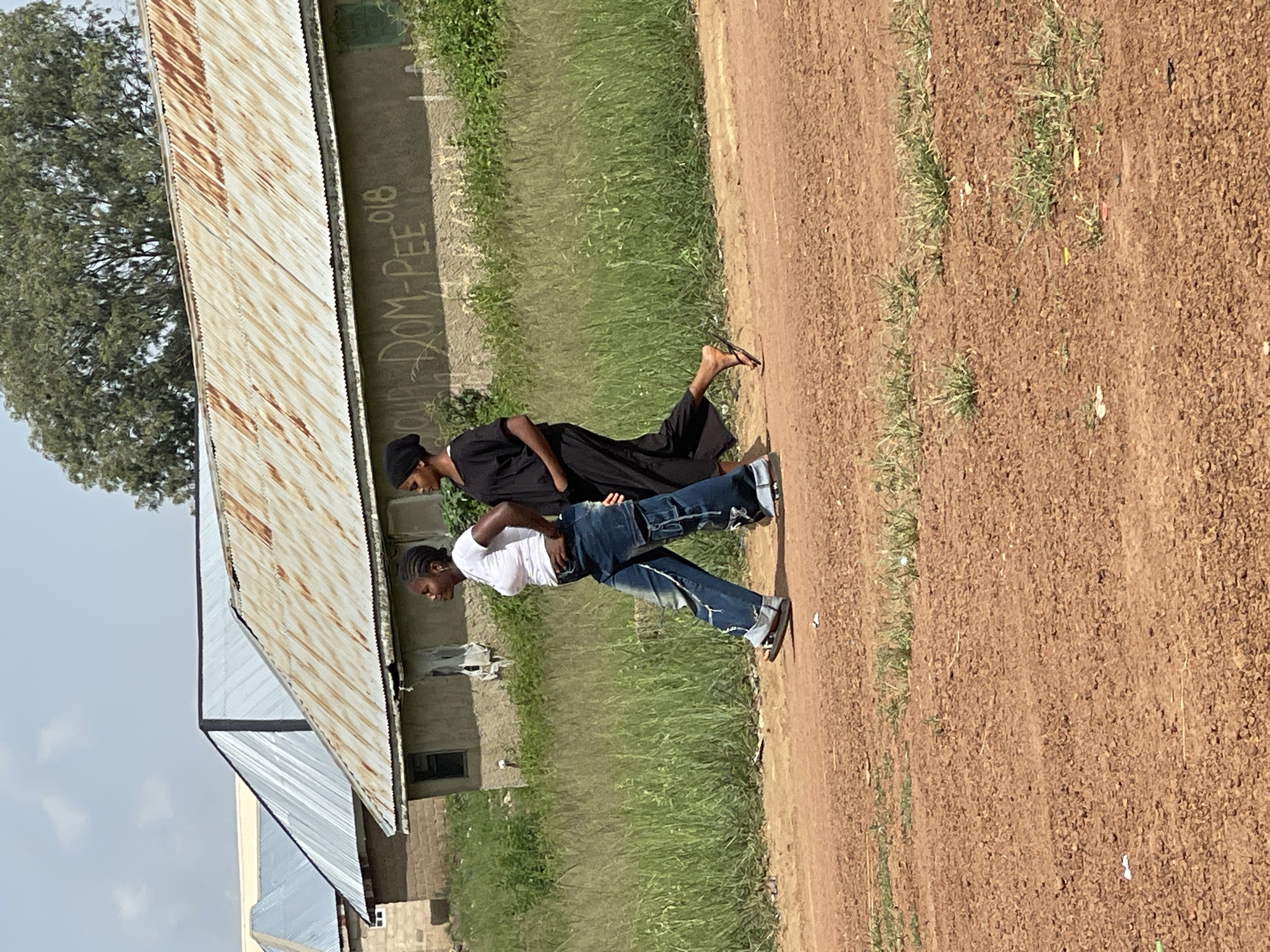 In Durumi, Nigeria, two young women walk side-by-side on a dirt road under a partly cloudy sky. The woman on the left wears a white t-shirt and distressed, baggy blue jeans, with her hands on her hips. The woman on the right is dressed in a black head covering and a flowing black garment. Behind them, a row of buildings with corrugated metal roofs are visible, one of which has graffiti on its wall that appears to read "OUR DOM-PEE 018". Tall green grass grows between the road and the buildings, suggesting a rural or semi-rural setting within the city.