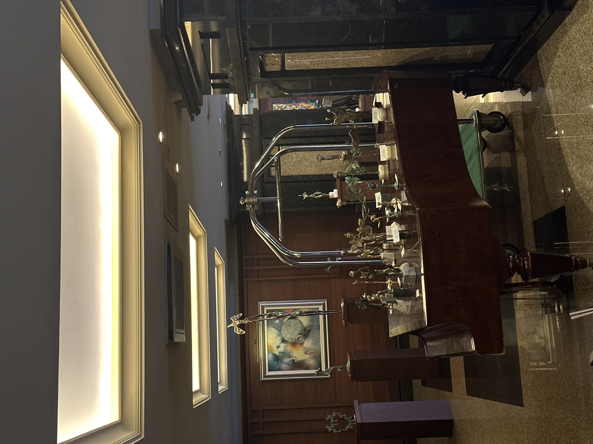 The image shows the interior of a building in Sofia, Bulgaria, featuring a grand piano adorned with numerous trophies. The piano is the focal point, its polished wood contrasting with the bright lighting emanating from large, recessed wall panels. Several smaller framed pictures are visible on the wall to the left. An elevator with a black marble façade is visible in the background. Trophies are arranged across the top of the piano.