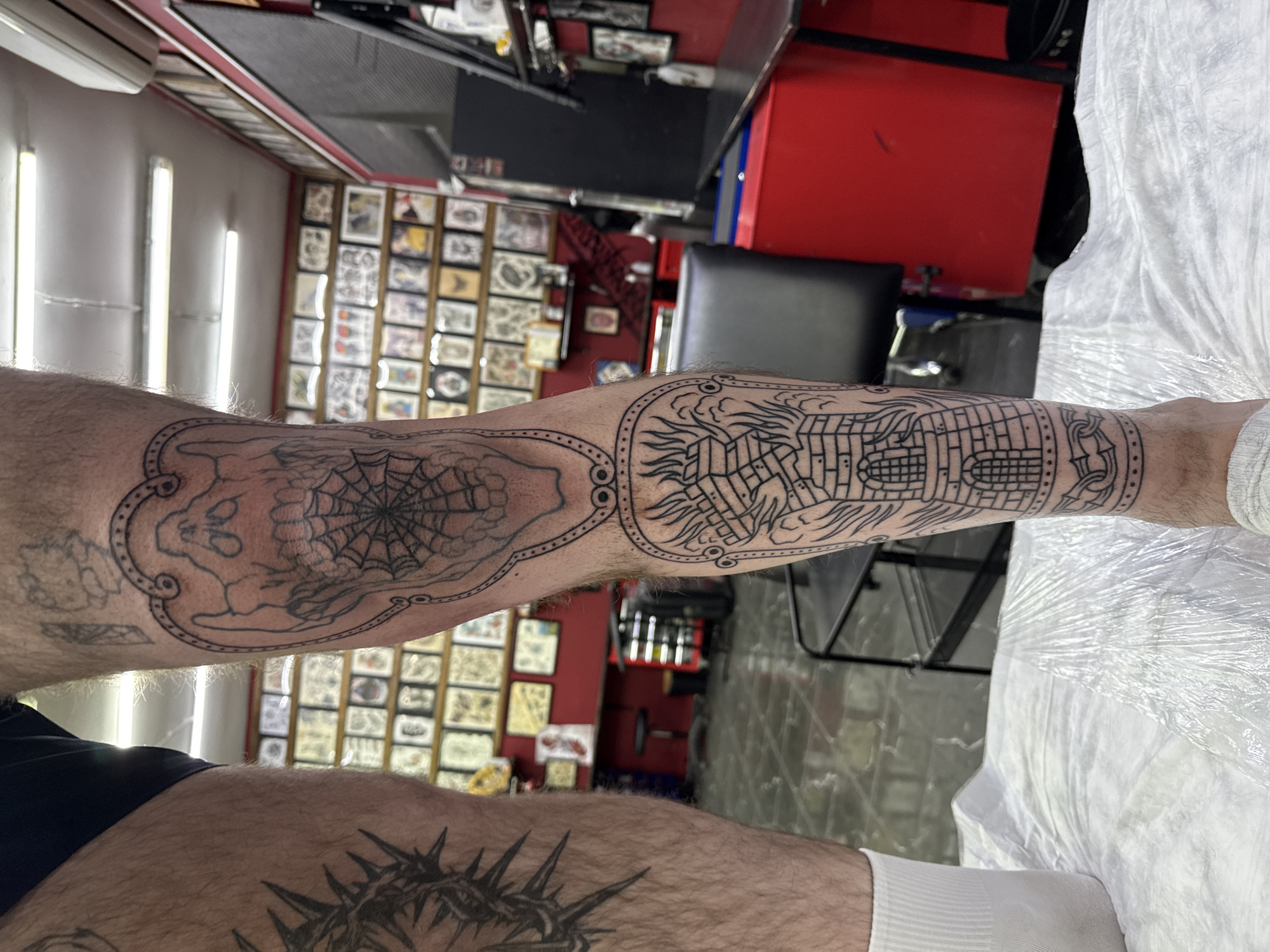 An image, captured from a low angle, shows a person's legs (likely a man's, given the hairiness) positioned within what appears to be a tattoo studio in Moscow, Russia. The individual is lying down, with their right leg extended towards the viewer, resting on a white, plastic-covered surface that suggests a tattooing bed or chair. A white sock is visible on the right foot.
The primary focus is on the intricate black-outlined tattoos on the person's right calf. These appear to be fresh outlines, uncolored. Two main designs are framed by a decorative border: on the left, a skull or skeletal face with a prominent spiderweb; on the right, a detailed structure resembling a castle or fortress, with flames or smoke emanating from its top. Further up on the left thigh, a portion of another tattoo depicting a crown of thorns is visible.
The background reveals typical elements of a tattoo parlor: a wall densely covered with framed tattoo flash art or completed designs, several fluorescent tube lights illuminating the space, and an air conditioning unit mounted on the wall. A red cabinet or storage unit and a dark chair are also visible, contributing to the utilitarian aesthetic of the studio. No specific time of day or emotions are discernible, but the setting indicates an ongoing or recently completed tattoo session. No discernible text is visible within the image.