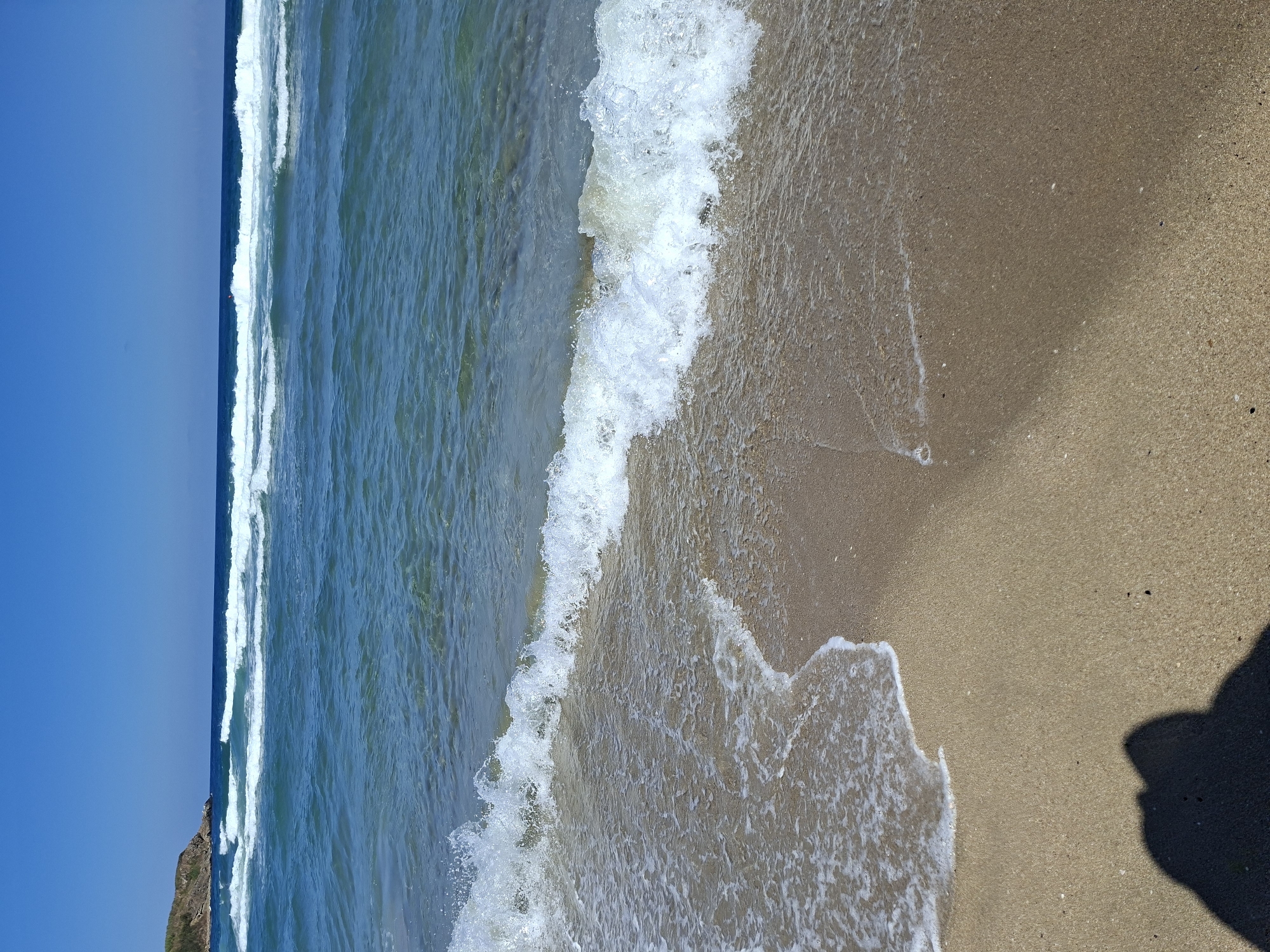 The image depicts a serene beach scene with clear blue water and a sandy shore. Gentle waves are seen rolling towards the beach, creating a frothy white edge as they reach the sand. In the background, a rocky outcrop covered in vegetation is visible on the left, and the horizon is marked by a clear blue sky. The foreground of the image shows the wet sand where the waves have receded, with a dark shadow of a person cast on the sand, indicating the presence of the photographer. The water is a vibrant turquoise, transitioning to deeper blue further out.