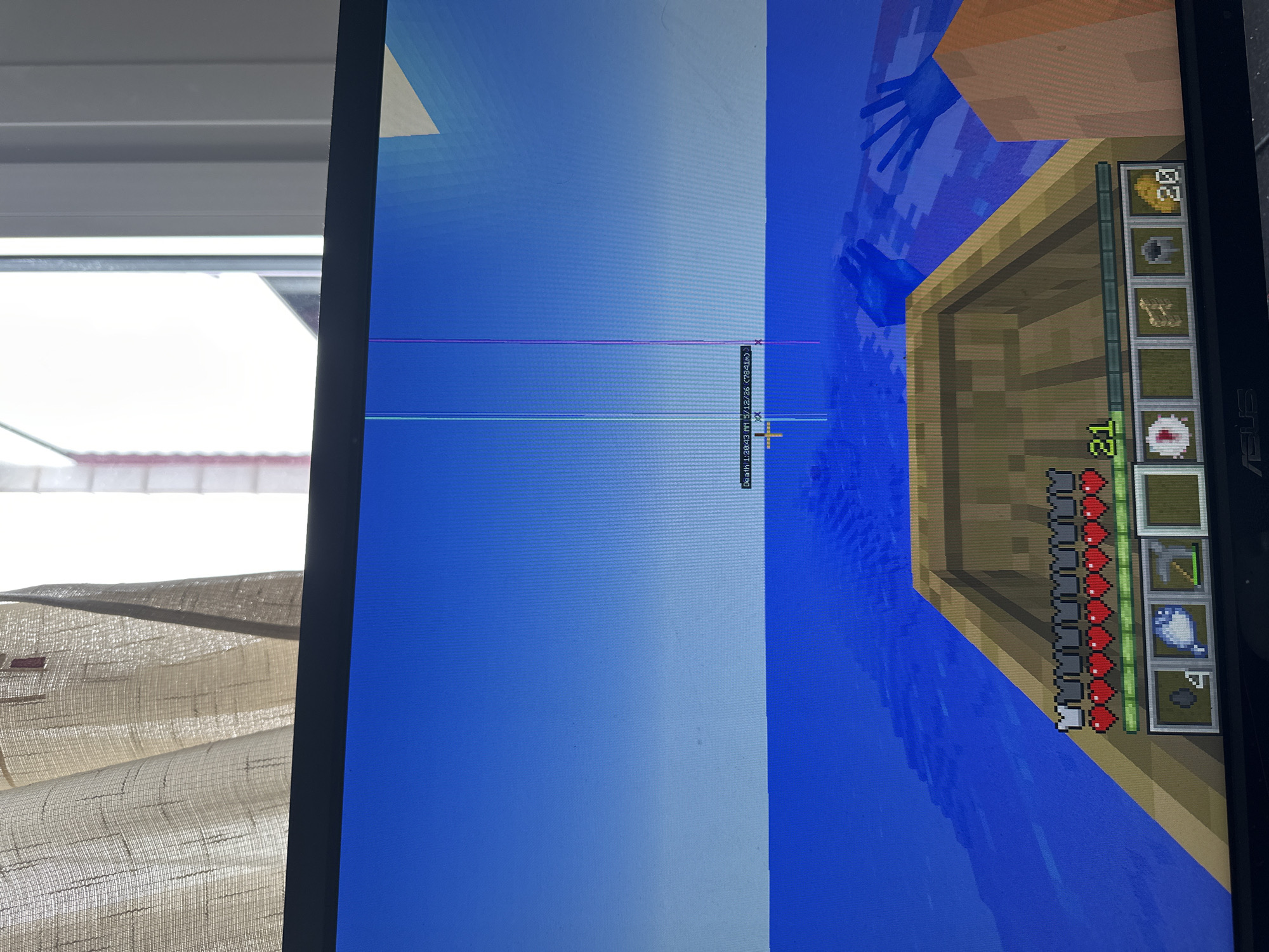 The image shows a close-up of a computer screen displaying gameplay from the video game Minecraft. The perspective is first-person, from inside a boat floating on water. The player's health bar and inventory are visible in the lower right. The game's debug information is visible on the screen, including coordinates. The environment outside the monitor is partially visible. A window with blinds is seen to the left of the screen.
The image provides no information about Bragança, Portugal.