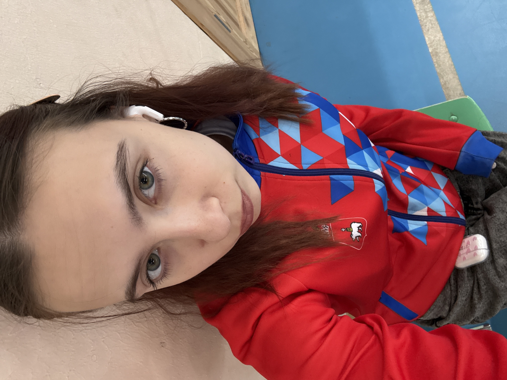 A young woman with light skin, hazel eyes, and brown hair is shown from a high-angle, close-up perspective. She wears wireless earbuds. Her red jacket, adorned with blue geometric shapes and a white emblem featuring an animal on the left breast, is visible. The background includes a light-colored wall and a blue and gray tiled floor. An object (likely a chair) with green edges is partially visible. The location is Berezniki, Russia.