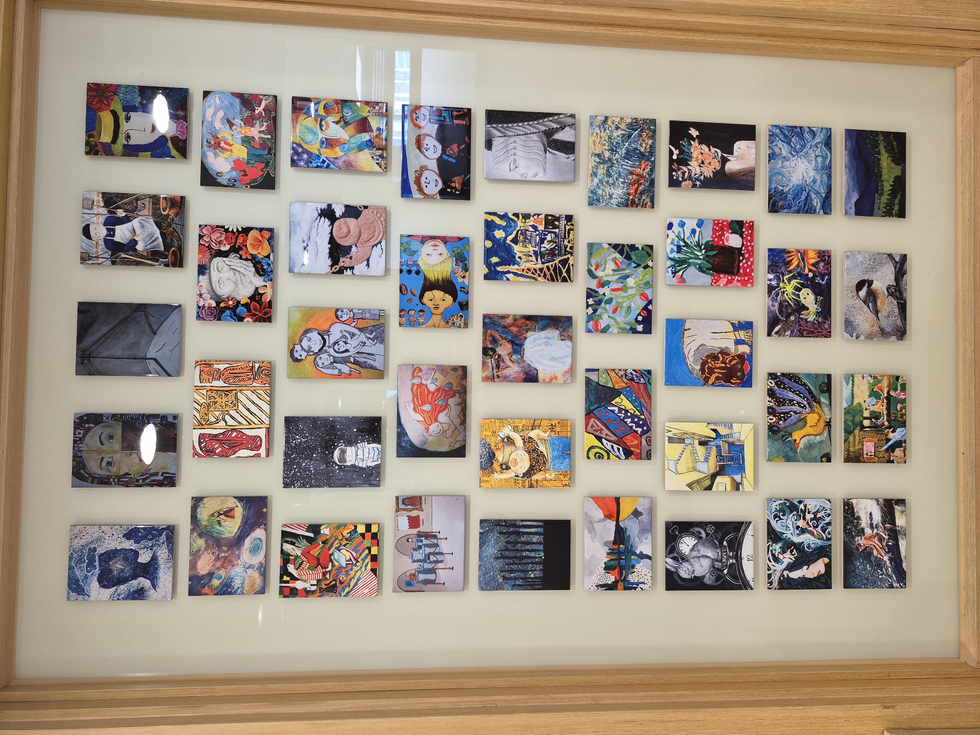 The image displays a framed collection of small, colorful paintings arranged in a grid on a light-colored wall. The paintings are of various sizes and subject matter, including portraits, landscapes, abstract designs, and whimsical scenes. Some of the paintings appear to be illustrations or artwork created by children. The frame is made of light-colored wood. The overall impression is of an art exhibition or a display of personal artwork. There are no discernible landmarks or elements within the image that would indicate a specific location in Seoul, South Korea. The scene is static, with no people or actions depicted.