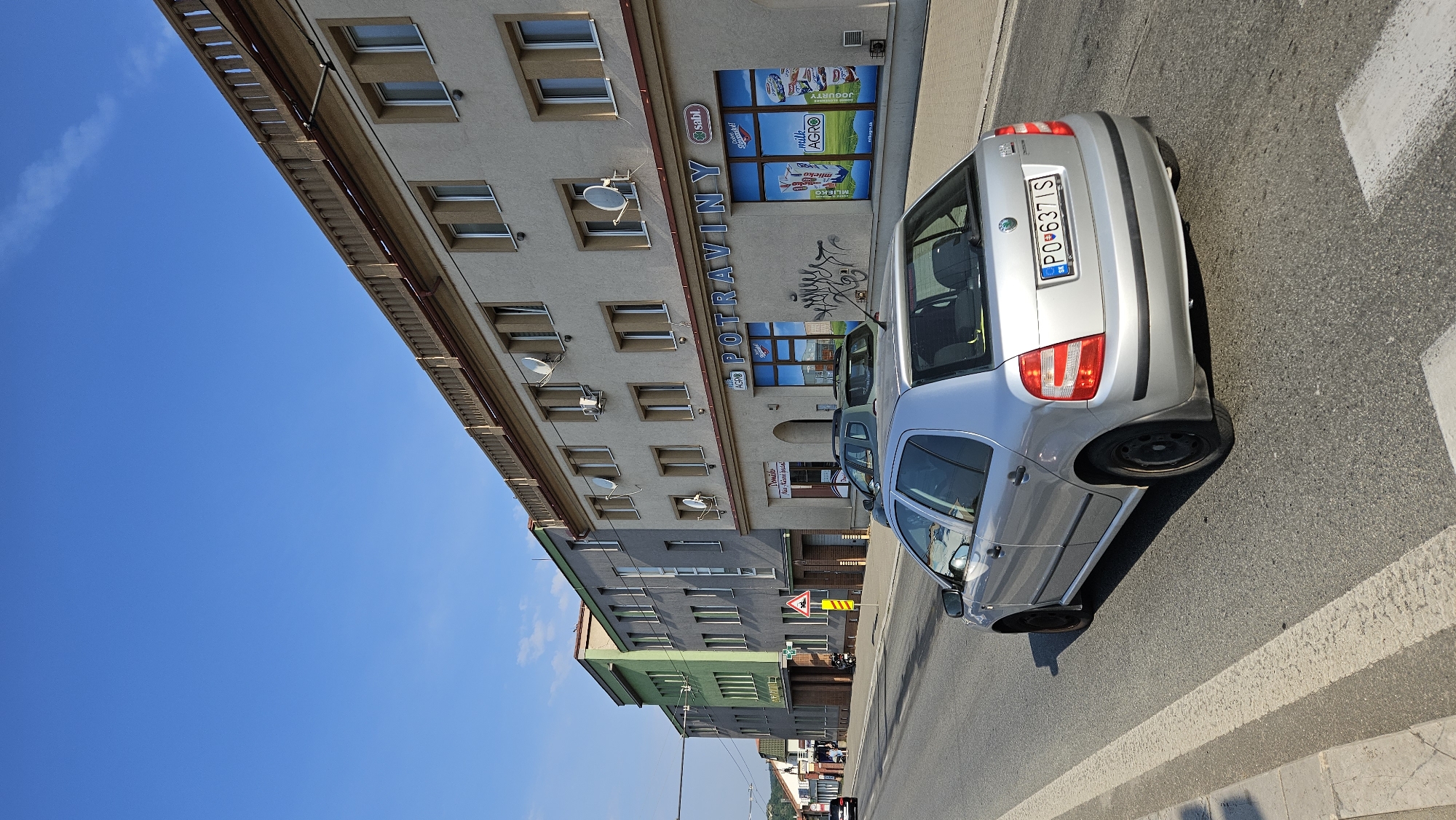 A silver Škoda Fabia is driving on a street in Prešov, Slovakia. The car is in the foreground, with its rear facing the camera. The street is asphalt and has white markings for a pedestrian crossing. The car has a license plate that reads "PO 637 IS".
In the background, there are several buildings lining the street. The closest building to the right is a light-colored, multi-story building with shops on the ground floor. One shop is labeled "POTRAVINY" (groceries). The building has multiple windows on the upper floors, some with satellite dishes attached.
To the left of this building is a grey multi-story building, and further to the left is a green multi-story building. These buildings also appear to have shops or offices on the ground floor and windows on the upper floors. A few people and a motorcycle are visible near the entrance of one of the buildings.
The sky is clear and blue, indicating a sunny day. The overall scene depicts a street in a European city on a bright day, with commercial buildings and traffic.