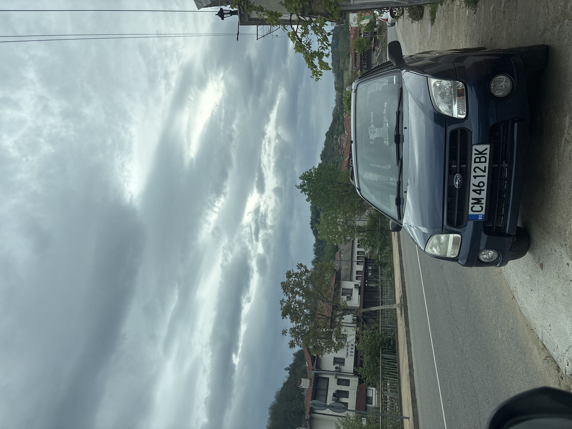 A medium shot of a blue Subaru car is parked on the side of a road. The car's license plate reads "CM 4612 BK". The sky is overcast with heavy clouds. Buildings are visible in the background, identifying the location as Zlatograd, Bulgaria. Vegetation is present along the roadside and in the background.