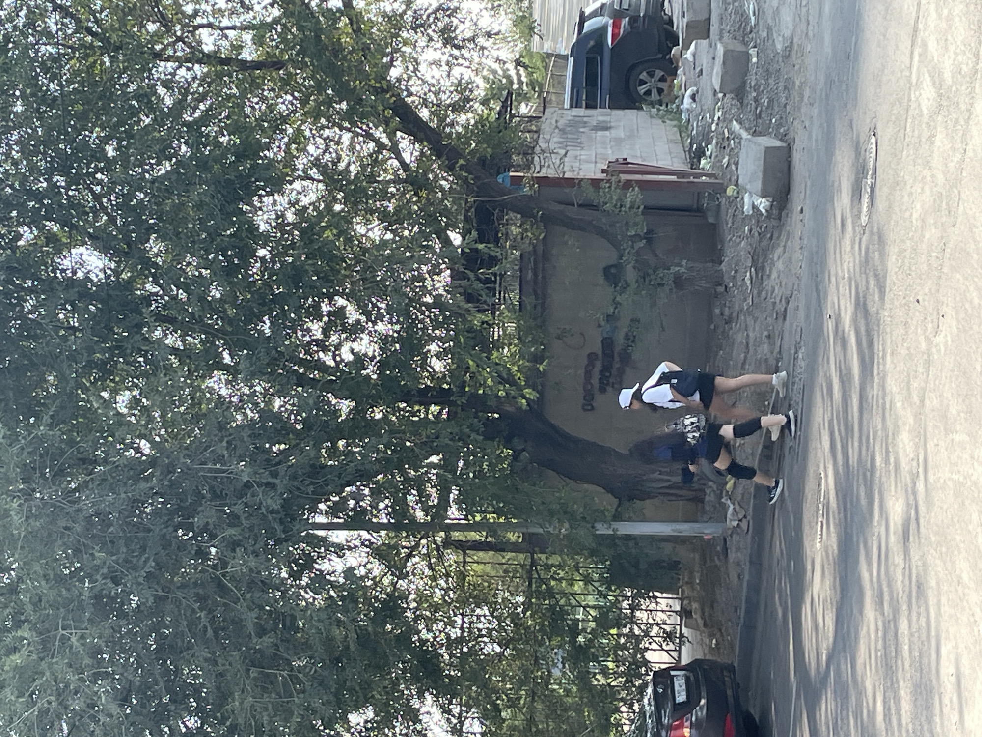 The image depicts two young women walking on a paved street in Almaty, Kazakhstan, under a large tree. The woman on the left has long dark hair and wears a dark blue long-sleeved shirt, black shorts, black knee pads, and white sneakers. She carries a dark bag. The woman on the right has dark hair and wears a white t-shirt, black shorts, and a white baseball cap. She has a white backpack. They are walking away from the camera towards the right side of the image.
The environment is somewhat dilapidated. Behind the women, a concrete wall with graffiti is visible, and there is a metal fence with greenery behind it. The ground on the right side of the street is littered with debris, including plastic bags and concrete blocks. To the far right, two dark-colored vehicles are parked, one is a black SUV. The street itself is asphalt with some markings and a manhole cover. Sunlight filters through the leaves of the large tree, casting dappled shadows on the ground. The overall impression is an urban scene with elements of disrepair.