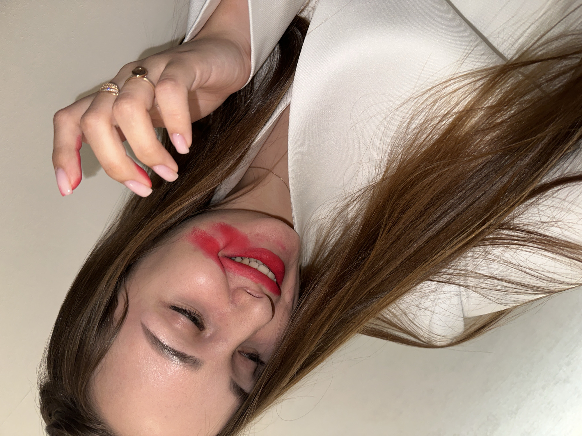 A woman with long brown hair is smiling with closed eyes. She is wearing a white blazer. Red lipstick is smeared around her mouth and also on her fingers. The background is a plain white wall. The photo is taken at an angle, making the subject appear tilted. Location information is unavailable.