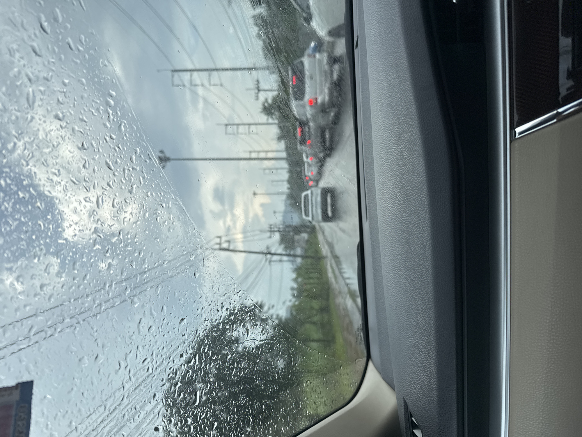 The image depicts a view from inside a vehicle looking through a rain-streaked windshield. Traffic is visible on a wet road, suggesting it is raining. Several cars are lined up in traffic, with their brake lights illuminated. Utility poles and power lines are visible along the side of the road. Lush green trees and vegetation are also present, hinting at a tropical environment. The sky appears overcast with clouds. The dashboard of the vehicle is visible in the foreground. The scene is consistent with being in a city like Kathu, Thailand, which experiences tropical weather and traffic congestion.