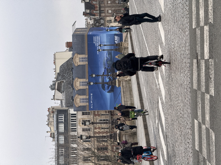 A Parisian scene unfolds with pedestrians and cyclists traversing a cobblestone street marked with zebra stripes. An Alpine advertisement hangs on a building, featuring an electric car model "4390". Architectural facades line the street, with winter trees visible. The sky is overcast.