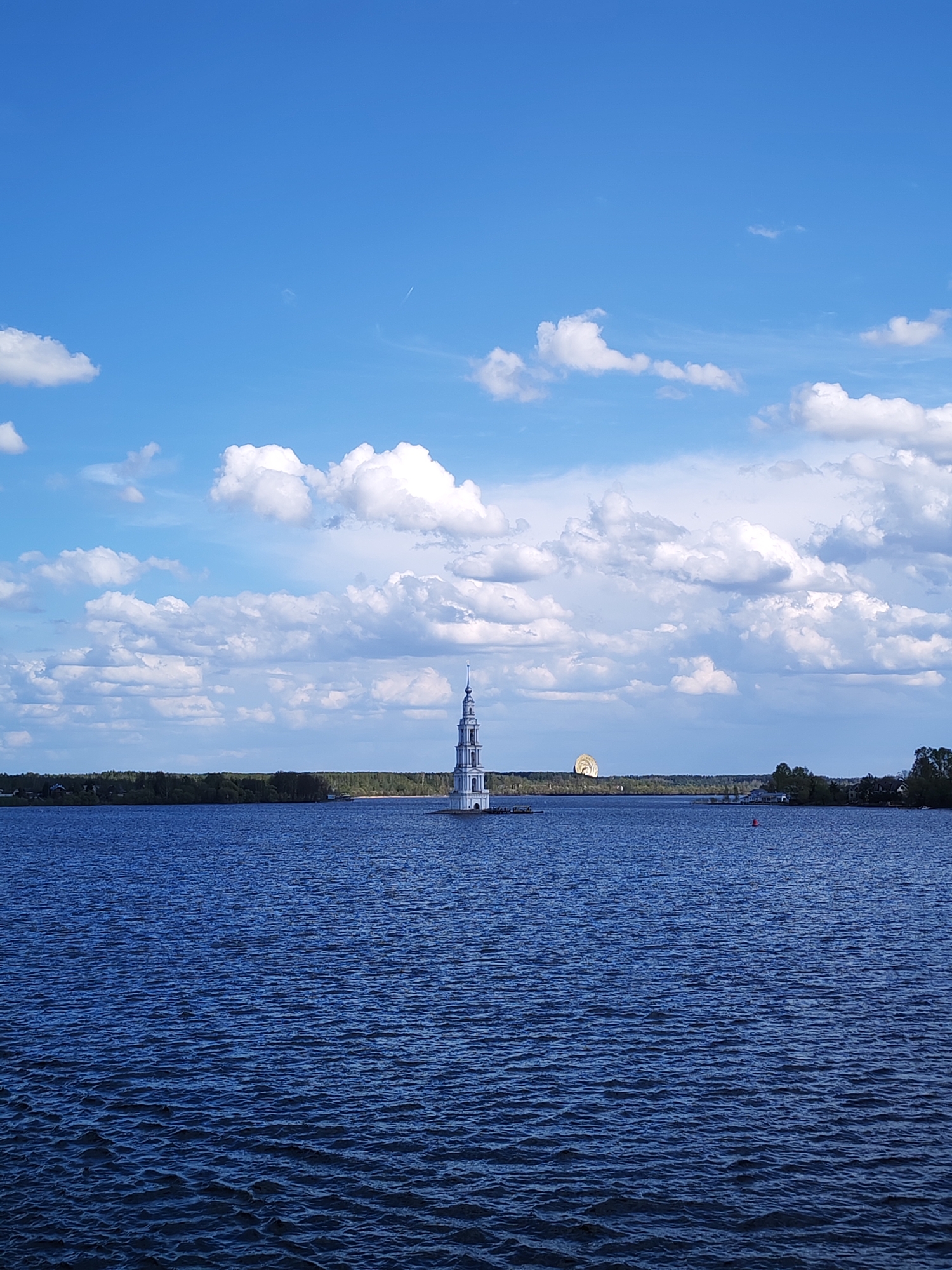 The image shows the flooded belfry of St. Nicholas Cathedral, a landmark in the city of Myshkin, Russia. The belfry stands in the middle of a body of water, with the shoreline visible in the background. The sky is blue with scattered white clouds. A domed structure is also visible on the shoreline.