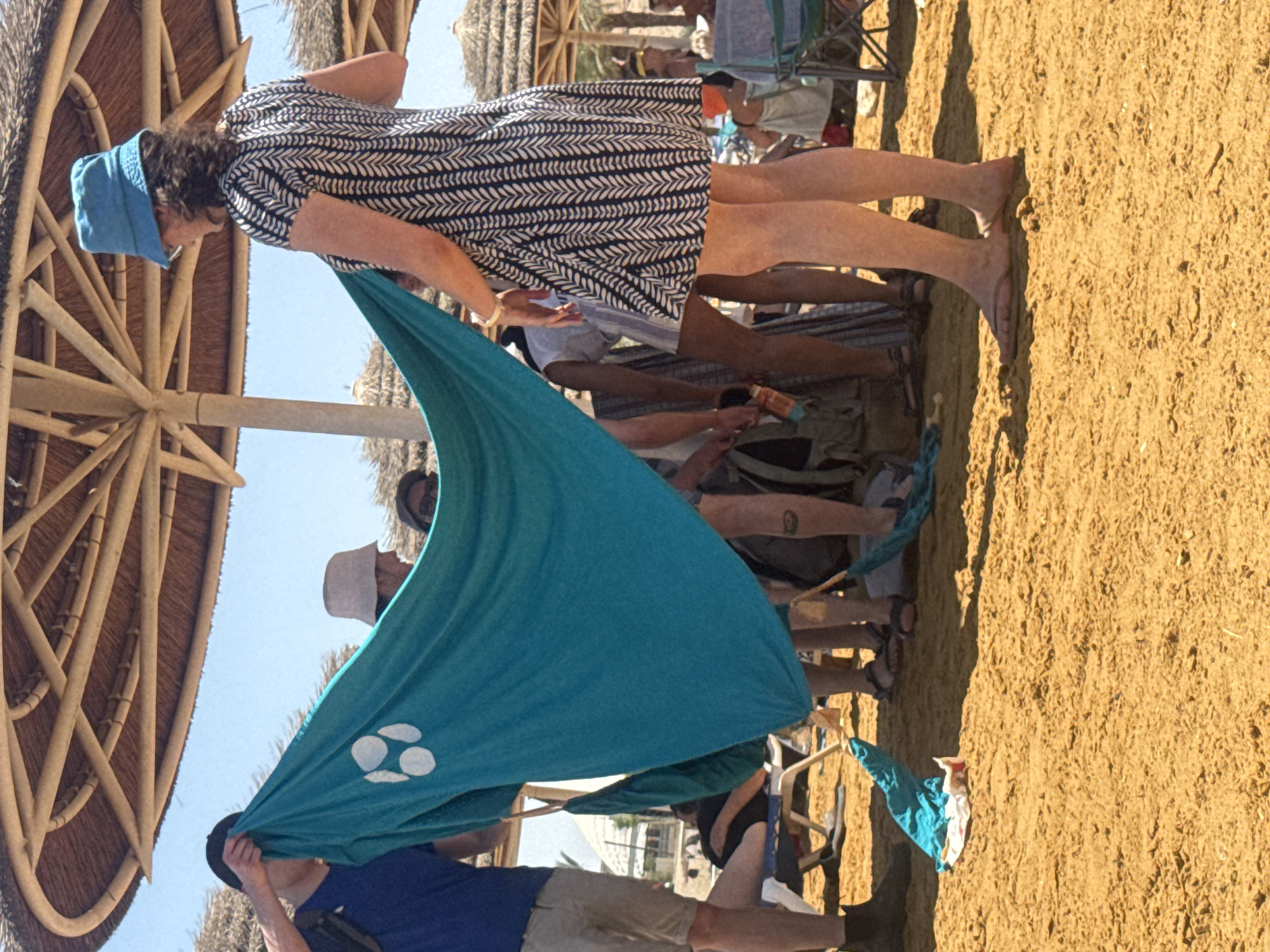 The image shows a beach scene in the Tamar Regional Council, Israel. A woman wearing a blue hat and a patterned dress holds up a large turquoise sheet or fabric with white circular designs. The sheet is also being held by other individuals. The group appears to be standing on a sandy beach, with several people and beach umbrellas visible in the background. Some individuals are seated in chairs.