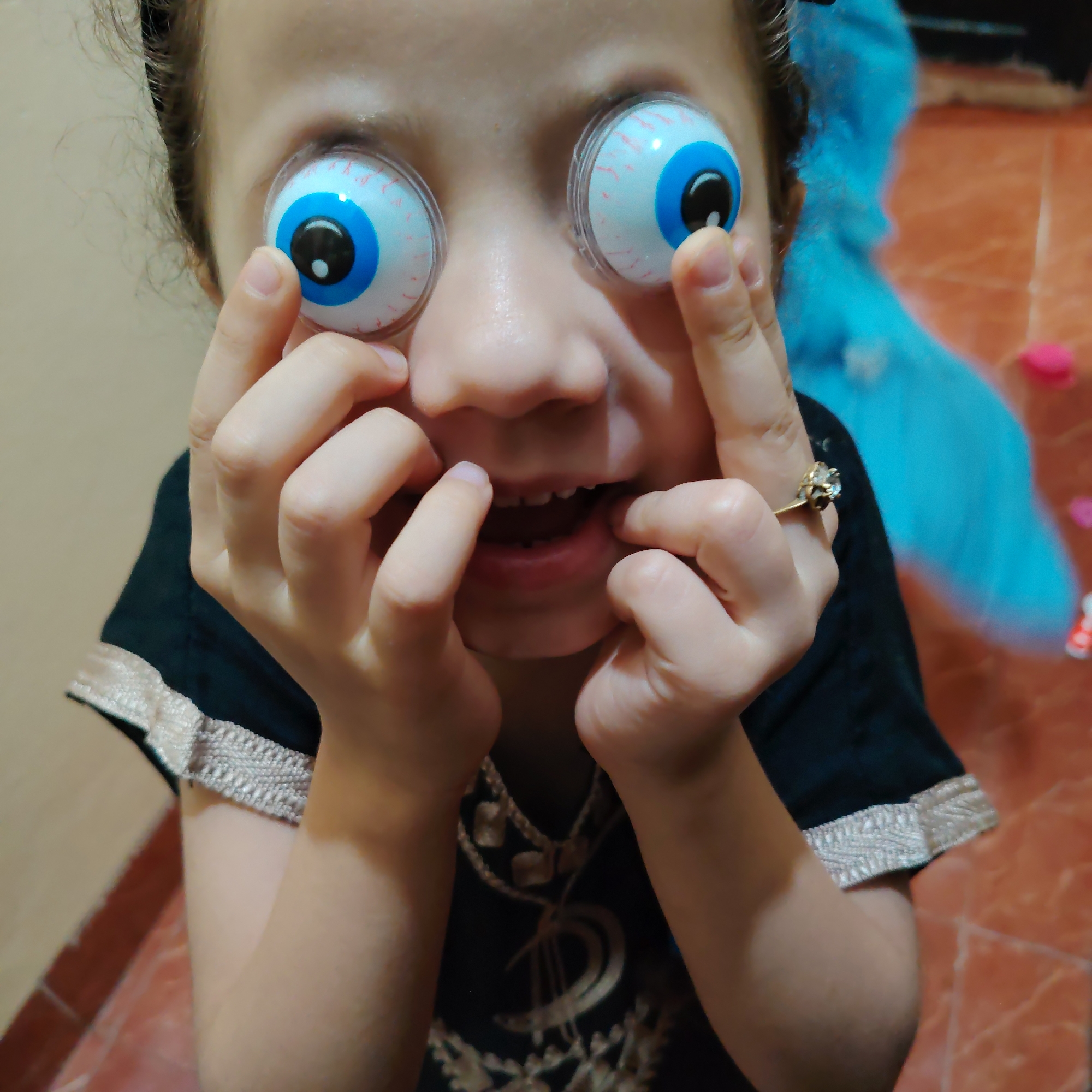In Ouaouizert, Morocco, a close-up shot shows a child wearing large novelty eyeballs over their own eyes. The child's hands are holding the fake eyeballs in place, with their fingers near their eyes and mouth. The child is wearing a dark garment with decorative trim. The background is blurred, showing a tiled floor and a blue fabric. The overall impression is one of playful expression and lightheartedness, with the child engaging directly with the camera.
