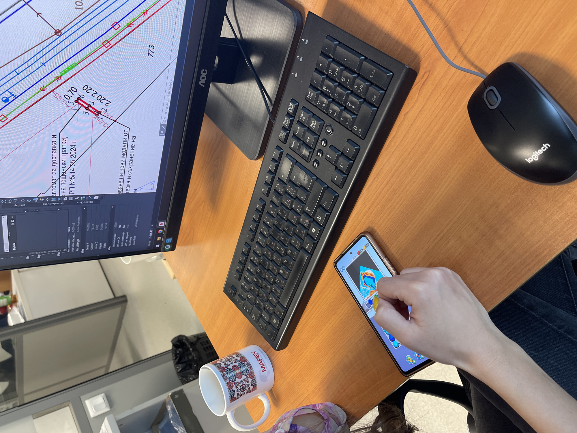 The image shows a desk scene in an office setting. A person is sitting at the desk, visible from the knees down in dark denim jeans and a hint of a floral slipper. A hand manipulates a game on a smartphone.
On the desk:
* A black monitor displays a technical drawing likely generated by CAD software.
* A black keyboard is next to the monitor.
* A black Logitech computer mouse is on the right side.
* The smartphone is next to the keyboard, displaying a game.
* A white coffee mug with "MAPEX" printed on it.
The environment includes a window with a view of an exterior, displaying architectural plans from the CAD software on the monitor. The location is most likely in Sofia, Bulgaria based on the text shown on the monitor.