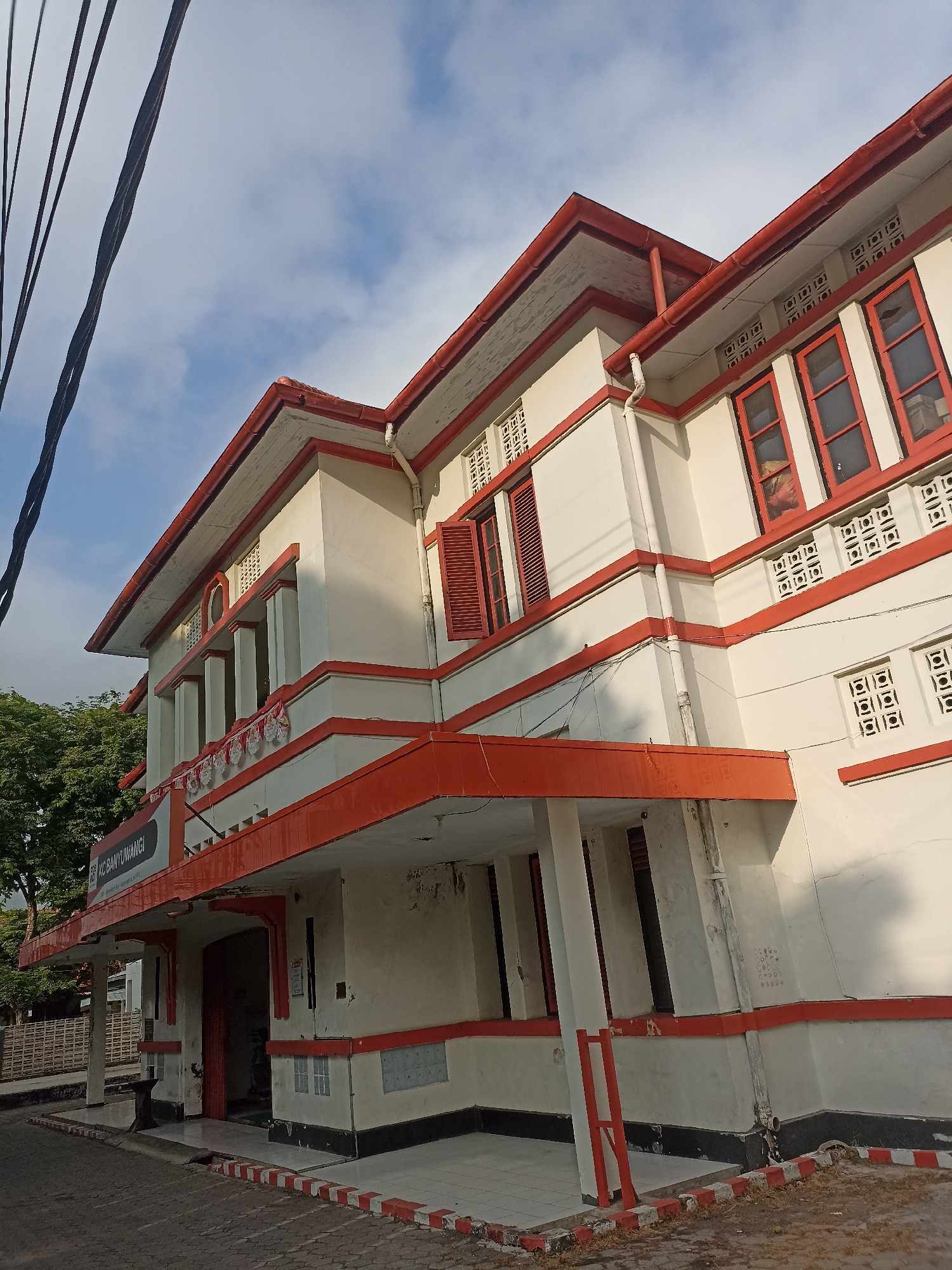 The image depicts the facade of a two-story building with distinctive red and white architectural elements, located in Kelurahan Kepatihan, Indonesia. The building appears to be a public or commercial establishment, possibly a bank, given the visible signage that reads "KC BANYUWANGI" on the left side. The structure features a prominent red-tiled roof with red trim along the eaves and edges. The walls are painted white, accented with red horizontal bands and decorative elements. There are numerous windows with red frames, some with louvered shutters. Ventilation bricks are also visible on the upper parts of the walls.
In the foreground, a paved walkway or driveway leads to the entrance of the building, which is partially obscured by a covered porch supported by pillars. A red and white striped border runs along the edge of the walkway. The environment is partially urban, with a few trees visible to the left and power lines crossing the top left of the frame against a blue sky with scattered clouds. No people are visible in the image. The overall scene suggests a bright, possibly sunny day.