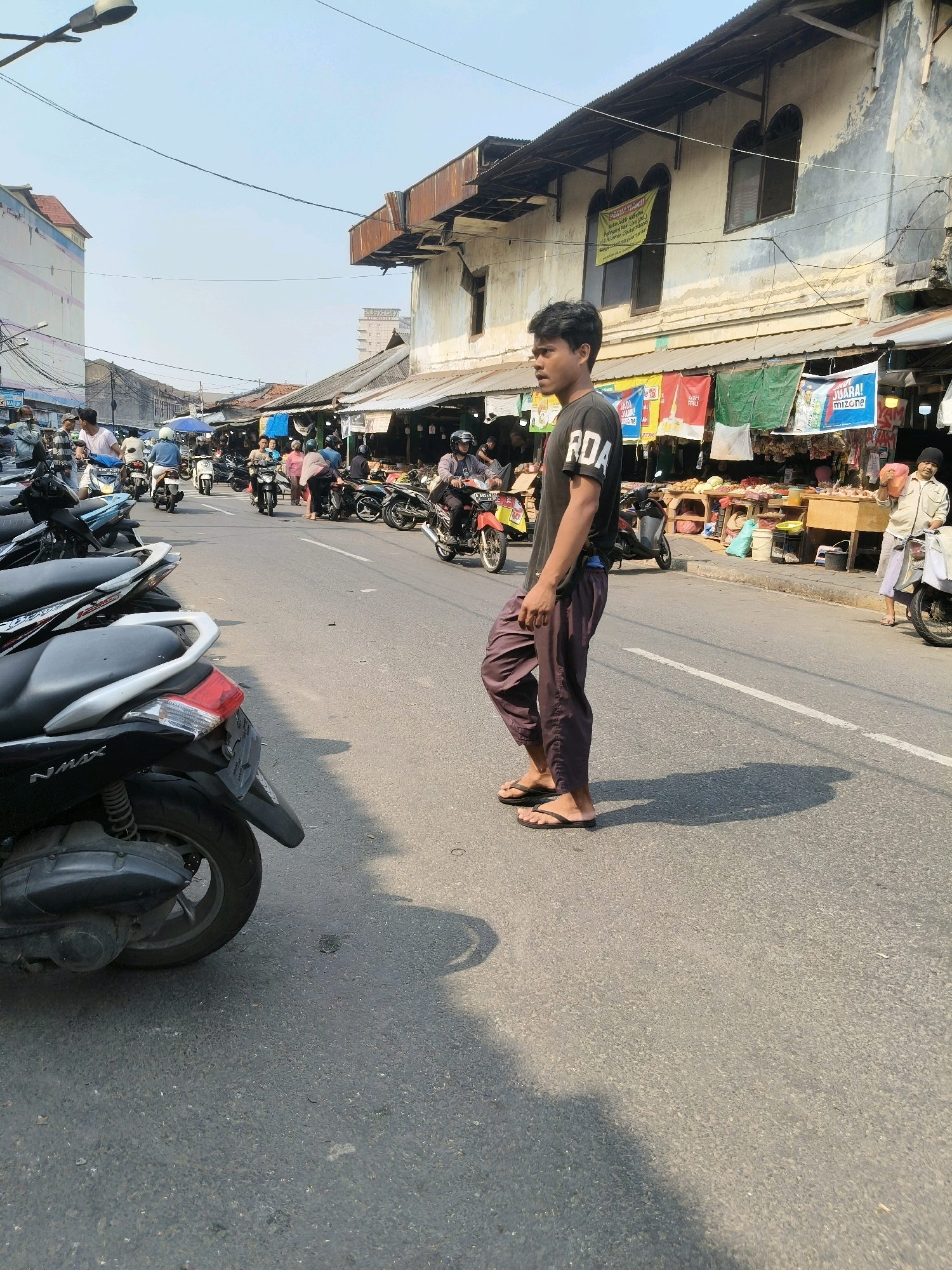 A young man stands on a paved road in South Tangerang, Indonesia, in front of market stalls. He is wearing a black t-shirt with white lettering that appears to read "RDA" and loose-fitting brown pants. He is also wearing dark flip-flops. The man is looking to his left. The road is lined with parked motorcycles on the left side of the frame and open-air market stalls on the right. Several people are visible in the background, some on motorcycles and others tending to the stalls. The stalls have various goods displayed, including food items. The buildings behind the stalls appear to be old and weathered, with some having corrugated metal roofs. The sky is clear and bright. The overall scene depicts a busy market area on a sunny day.