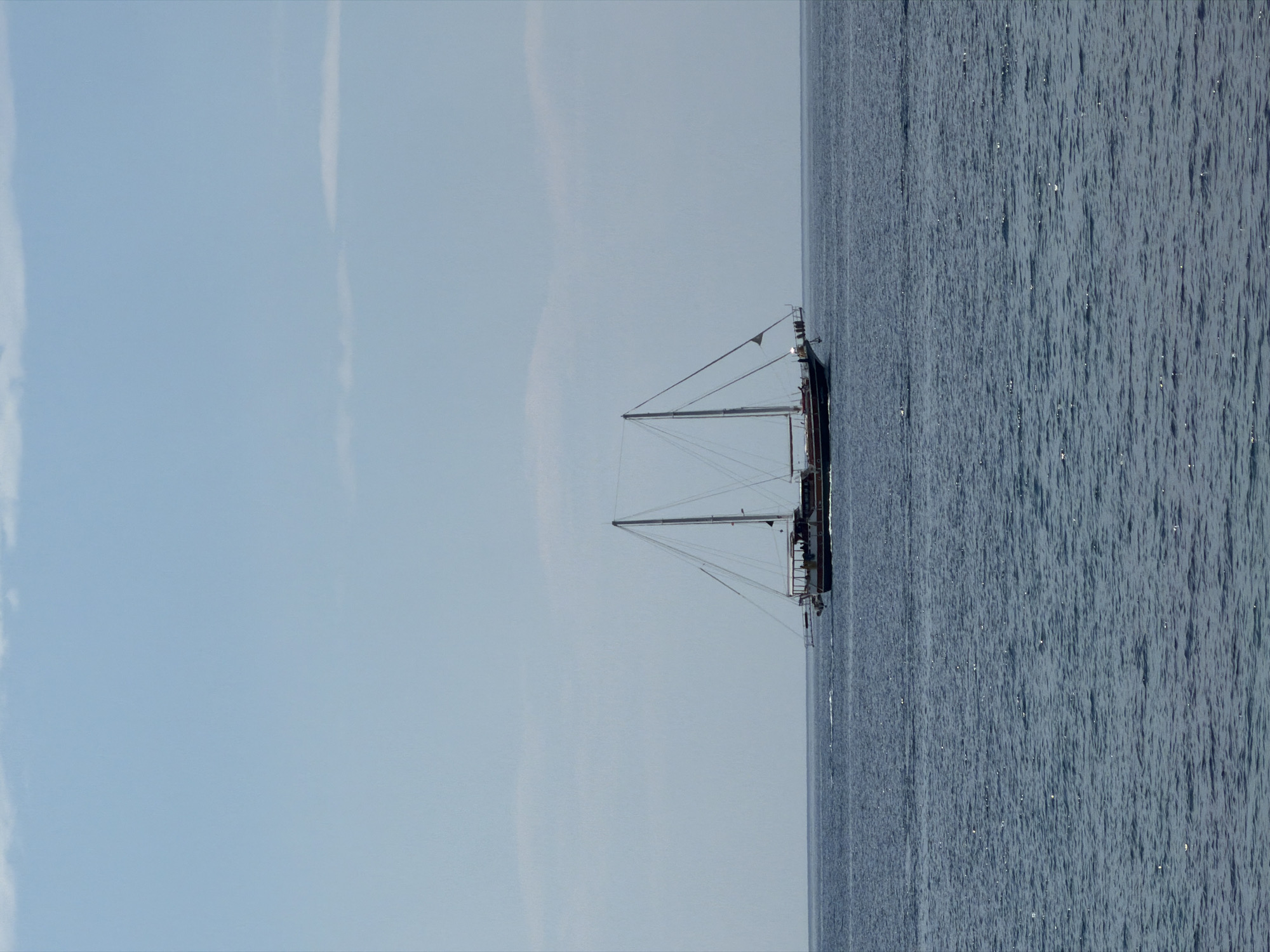 A long shot features a two-masted boat at sea, horizontally oriented. The right side of the image is occupied by the sea, its surface marked by subtle waves and a slight texture. To the left, the sky fills the remaining space, a light blue gradient with faint streaks of clouds. The boat is positioned roughly in the middle of the frame, its masts and rigging contrasting against the lighter sky. The location is Budva, Montenegro.