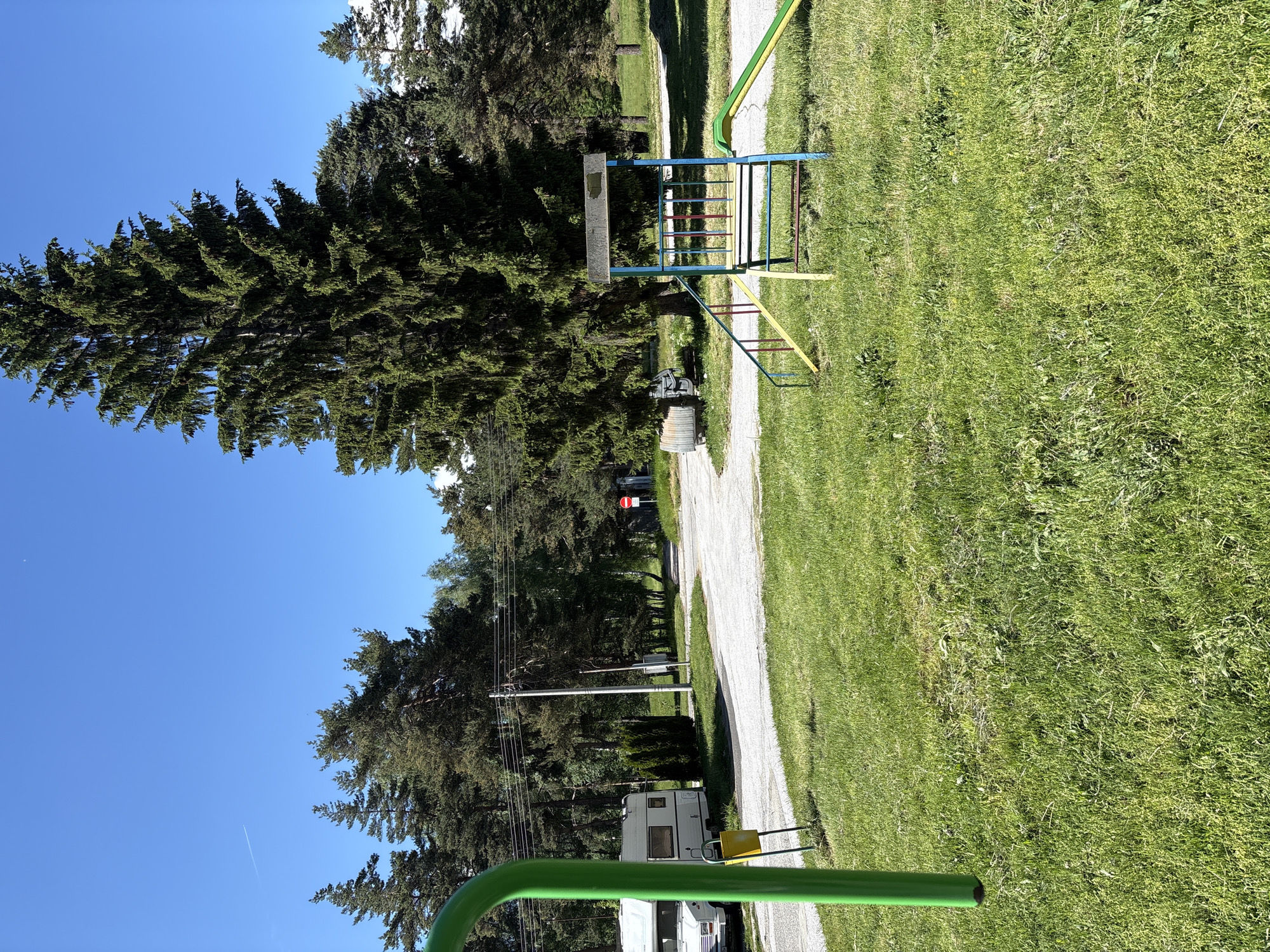 The image depicts a grassy park area in Panagyurski kolonii, Bulgaria, under a clear blue sky. A large evergreen tree stands prominently on the left. A gravel path runs through the center of the image, leading into the distance. A colorful playground structure, with blue, yellow, and red bars, is visible on the right side of the path. Further down the path, a white camper is parked. In the foreground, a curved green object is partially visible. Power lines run along the left side of the path. A no-entry sign is situated further along the path.