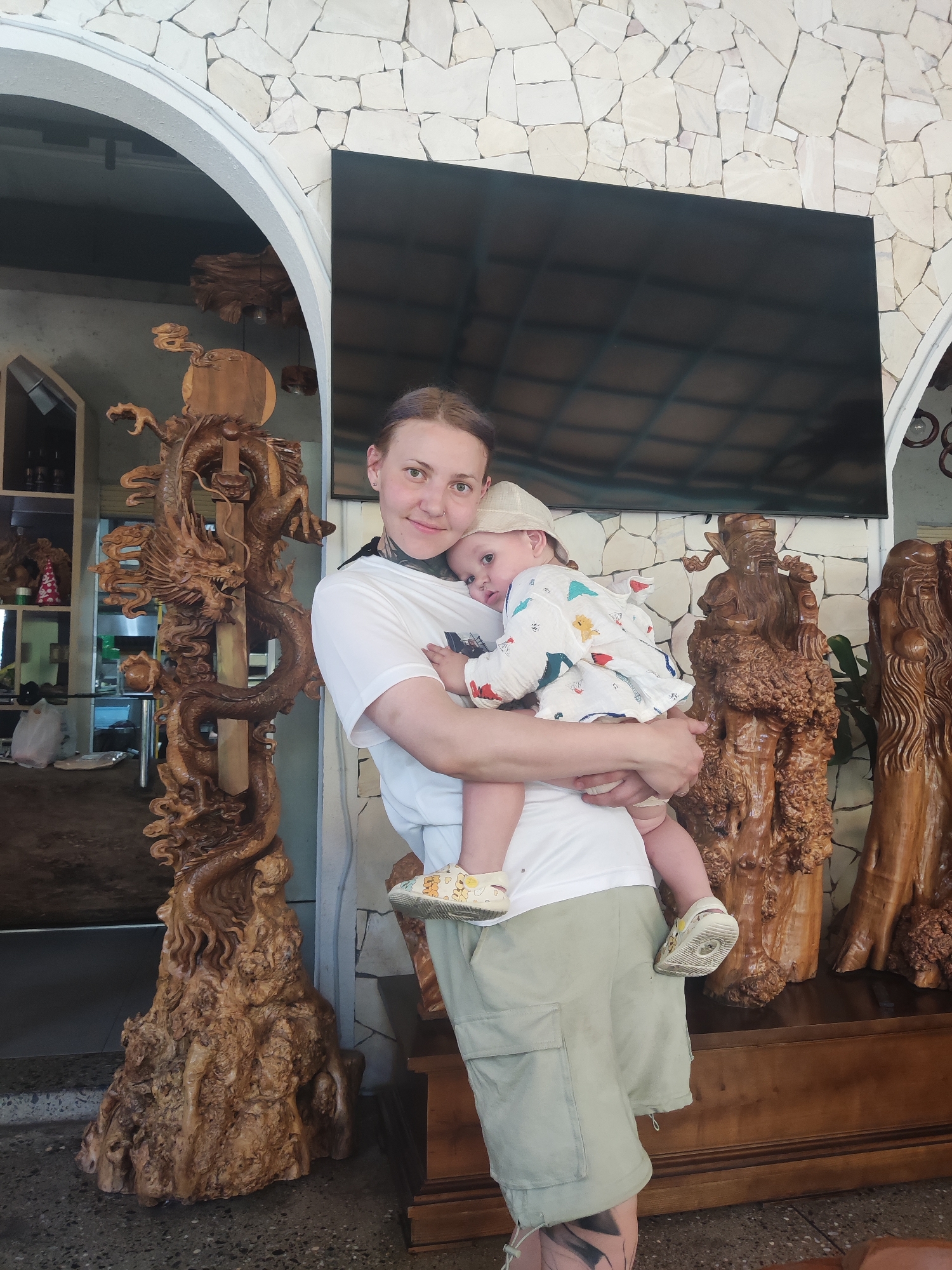 An indoor portrait features a woman holding a baby, likely taken in a shop or gallery specializing in wooden carvings in Đà Nẵng, Vietnam. The woman, with dark hair, a nose ring, and tattoos on her neck and lower leg, wears a white t-shirt and light green cargo shorts. She smiles subtly at the camera while the baby, wearing a patterned top, a light hat, and distinctive shoes, leans against her, looking away.
The background is filled with elaborate wooden sculptures. To the left, a tall, intricately carved wooden dragon spirals upwards, its base crafted from natural burl wood. To the right of the subjects, two large, dark wooden figures, resembling wise men or deities, stand on a wooden surface, also carved from what appears to be burl or natural wood forms.
The setting includes a distinctive wall of irregularly shaped, light-colored stones, with white archways framing parts of the scene. A large, dark rectangular screen or television is mounted above the wooden figures, reflecting the ceiling. Through an archway on the far left, shelves with various items are visible. The floor is concrete. The lighting suggests it is daytime. There is no discernible text in the image.