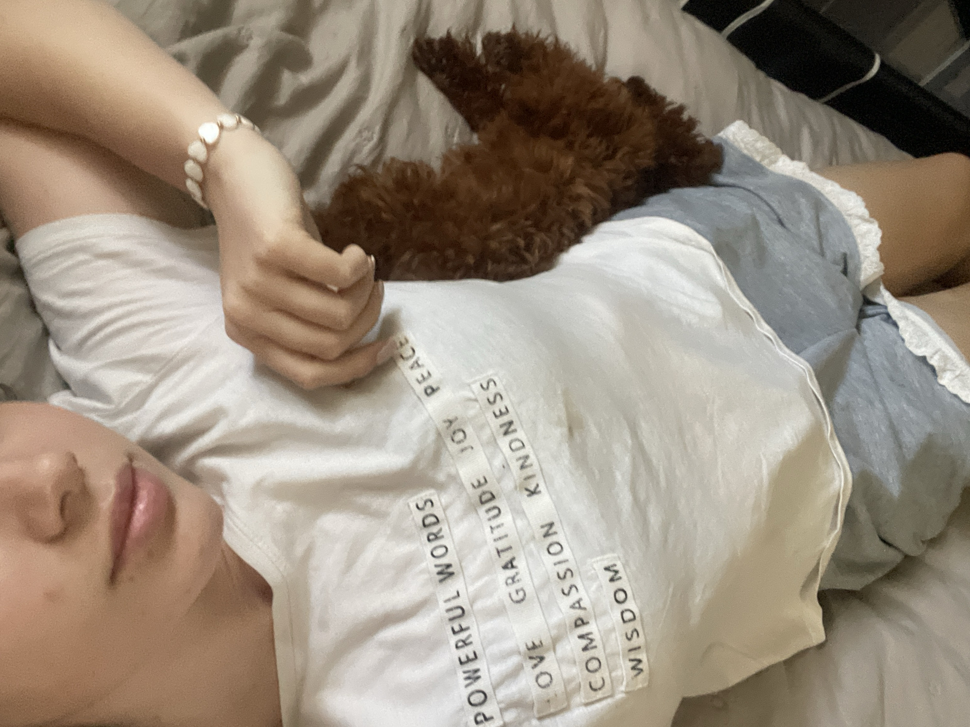 A person lies on a bed with a brown poodle resting on their chest. The person is wearing a white t-shirt with the words "POWERFUL WORDS, LOVE, COMPASSION, WISDOM, GRATITUDE, JOY, PEACE, KINDNESS" printed on it. They are also wearing light blue shorts with a ruffled edge. The person has a bracelet on their wrist. The bed has a gray duvet. The location appears to be indoors, likely a home, but there is no specific information suggesting Sofia, Bulgaria.