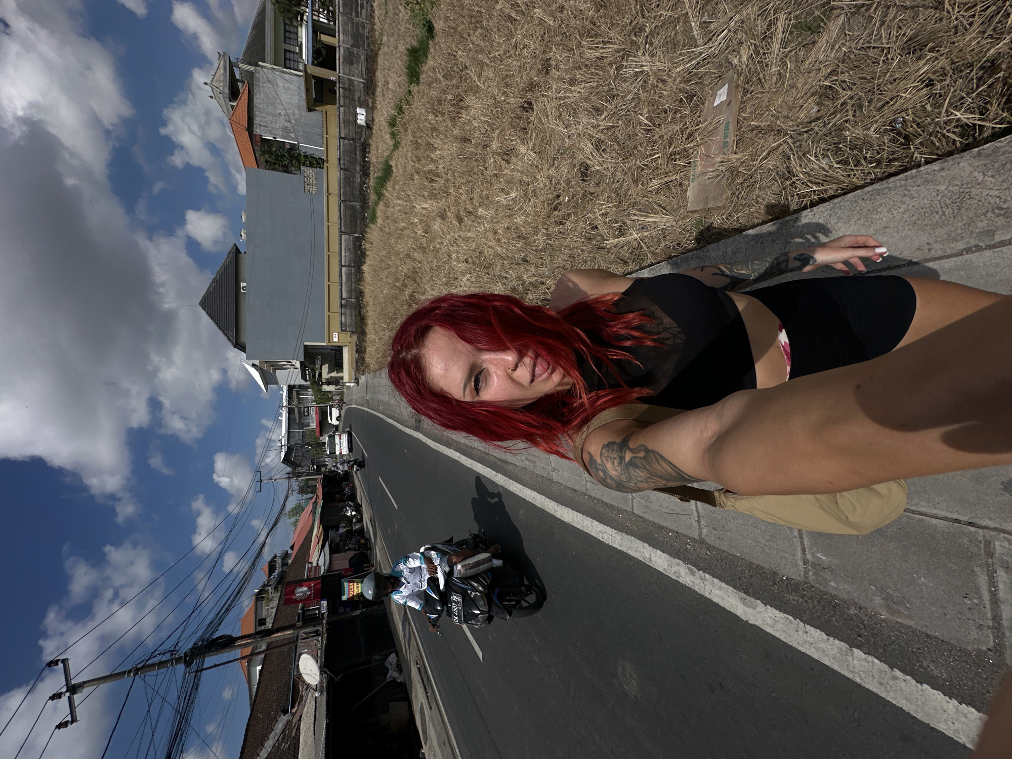 A young woman with bright red hair, wearing a black crop top and black shorts, is taking a selfie while walking on a sidewalk next to a street in North Kuta, Indonesia. She has tattoos on her arm. The street is paved and has white lane markings. A person wearing a helmet and a white shirt is riding a scooter on the street. The sidewalk is made of concrete. To the right of the sidewalk is a dry, grassy field. In the background, there are buildings, power lines, and a cloudy blue sky. The sun is shining, casting shadows.